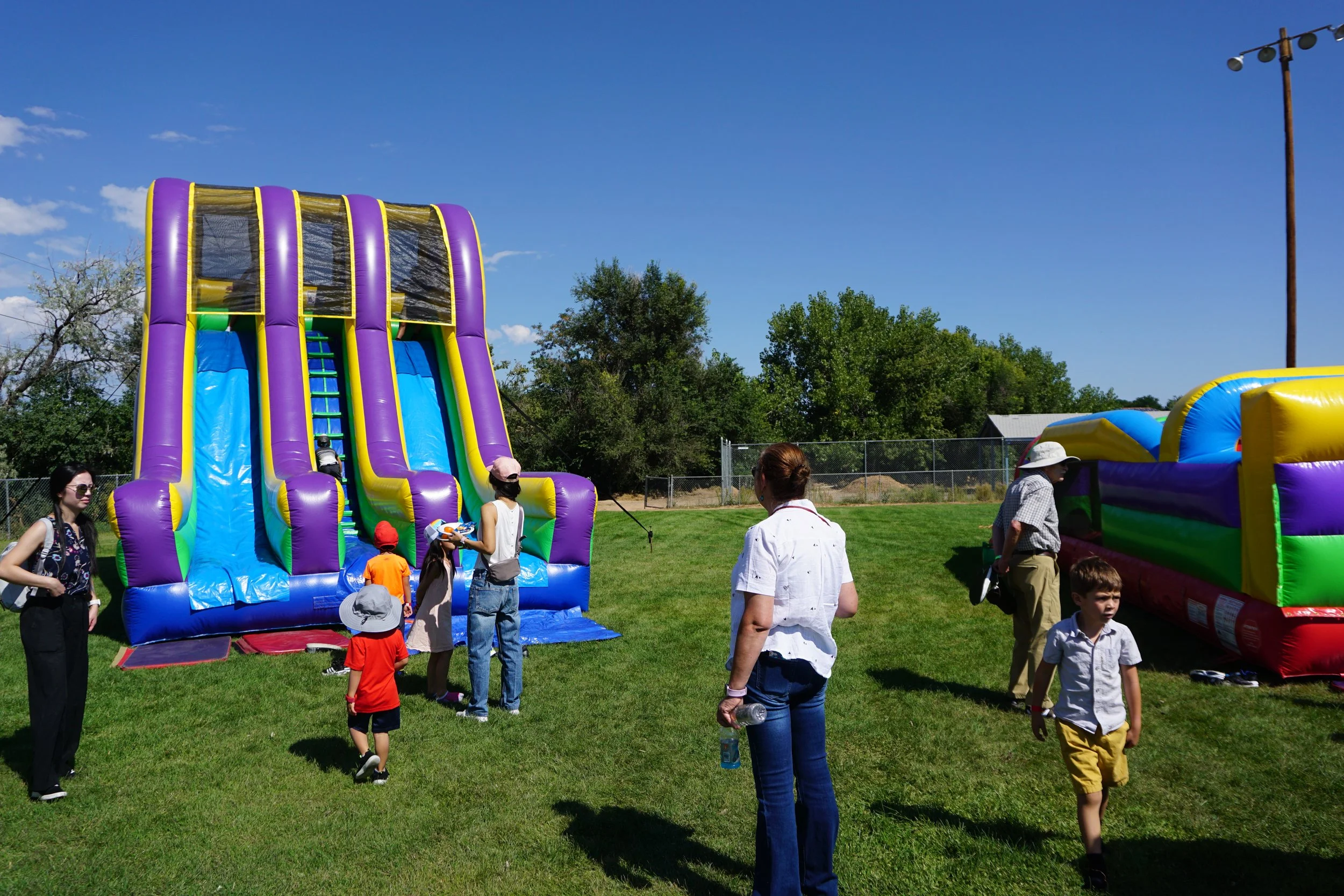 People, including children and adults, gathered on a grassy field with inflatable slides and bounce houses at a fair or festival on a sunny day.