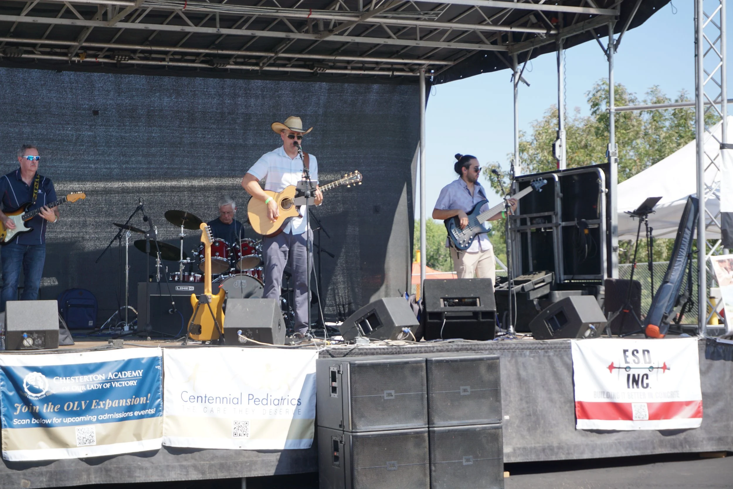 A band performing on an outdoor stage during daytime, with musicians playing guitars and drums. The stage has a black backdrop and several speakers in front.