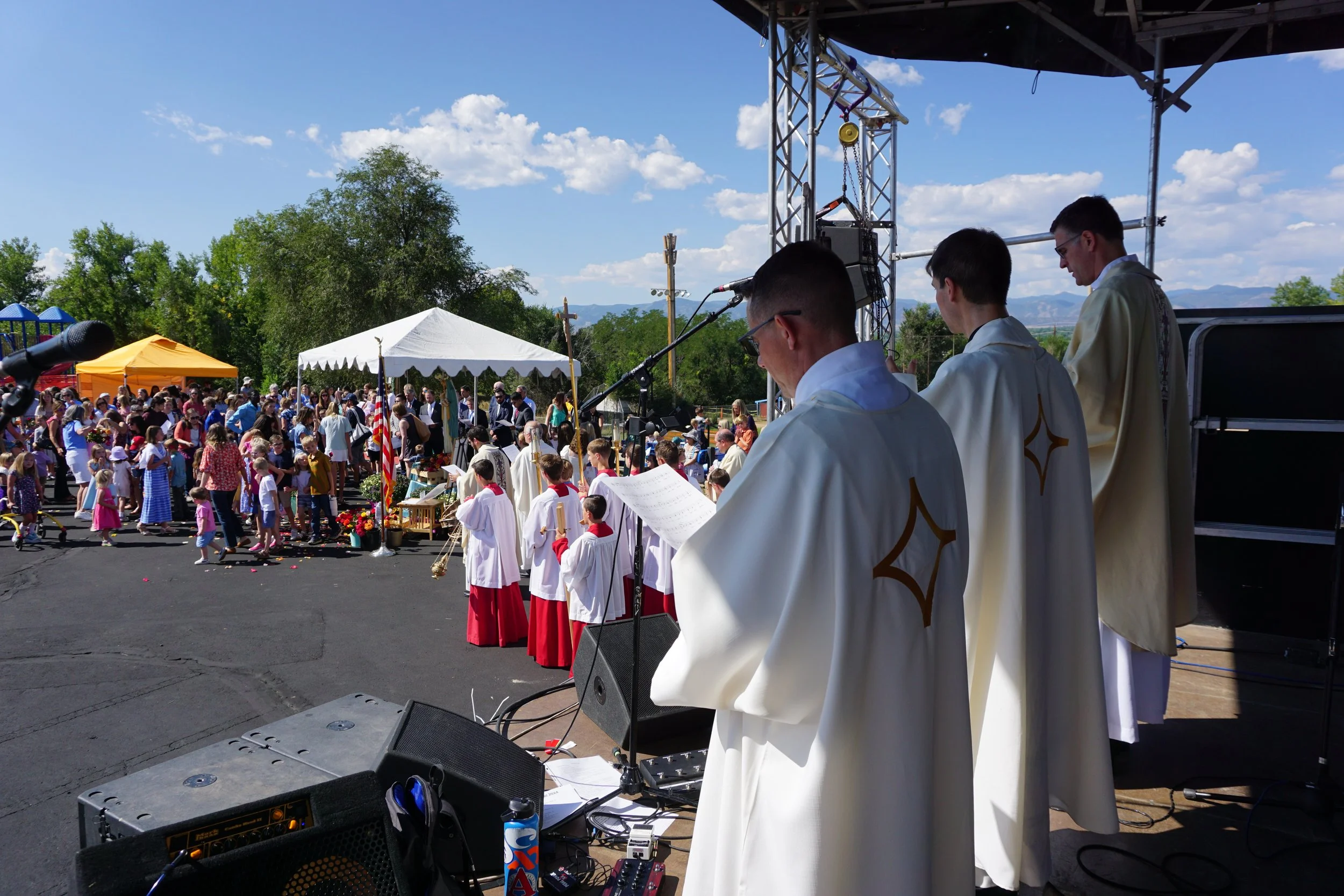 Clergy members performing a religious service outdoors with a large congregation, during a sunny day with blue sky and clouds.