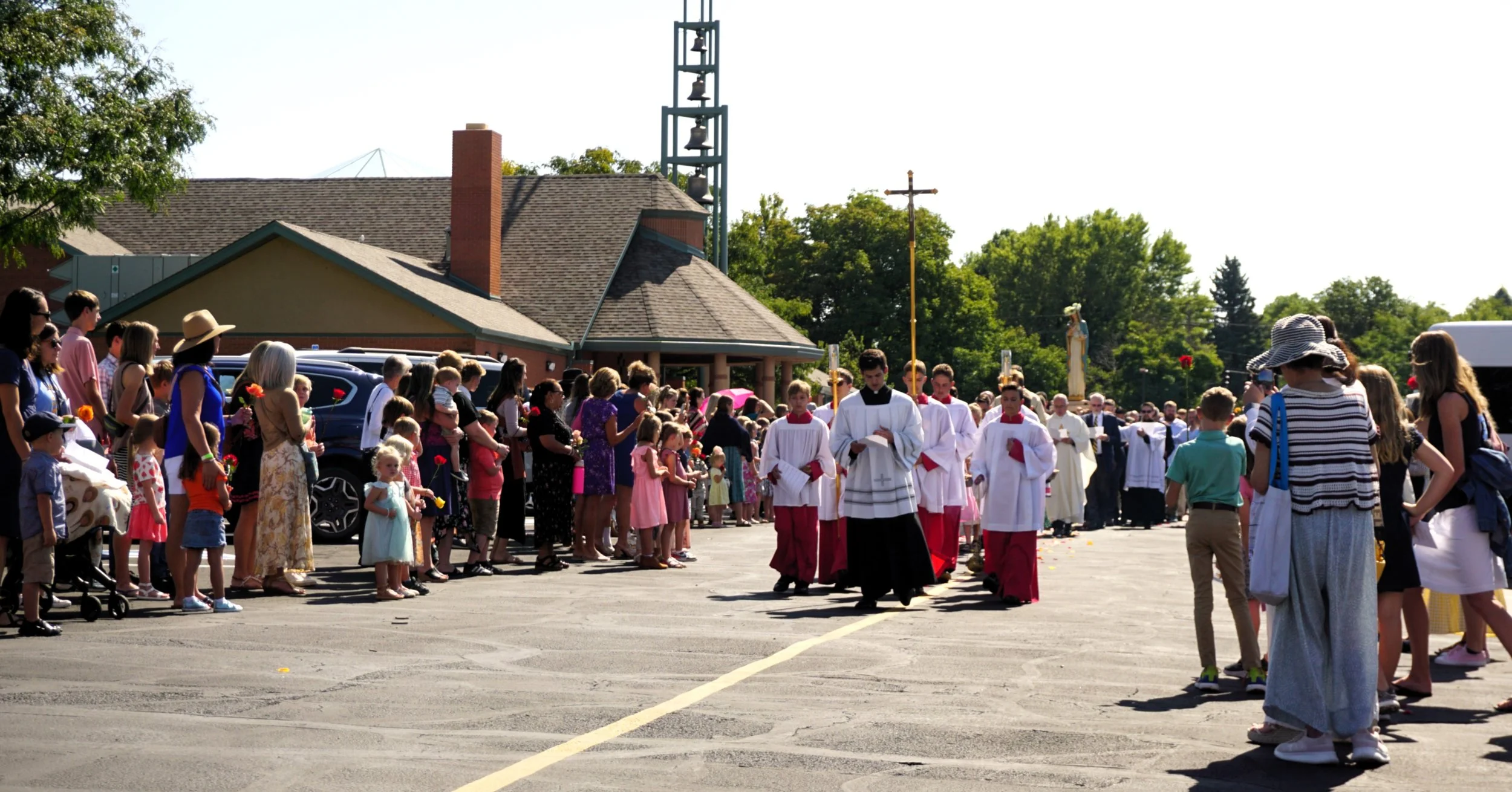 A religious outdoor procession with clergy members in white vestments and people holding flowers, gathered outside a church building on a sunny day.