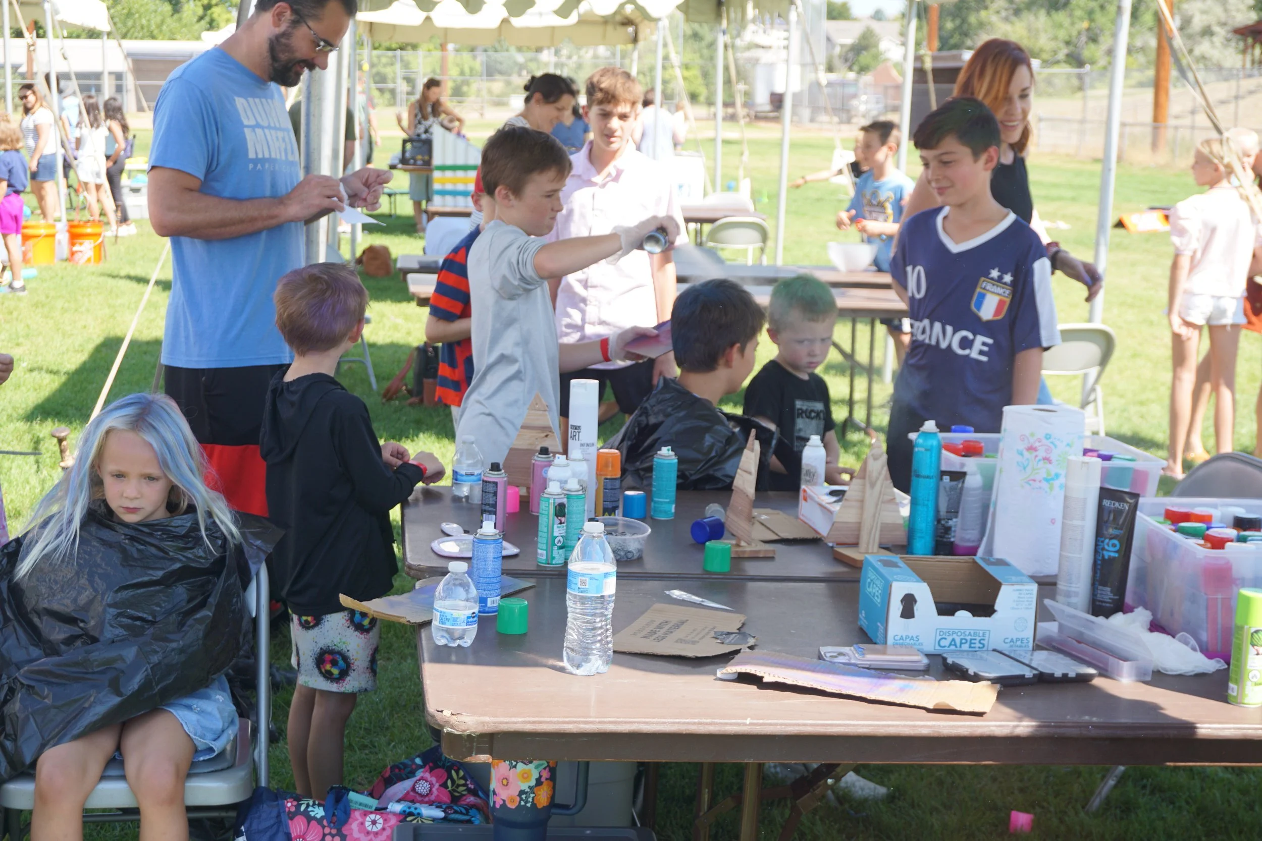 Children and adults at a community outdoor event with tables of arts and crafts supplies, tents, and green grass.