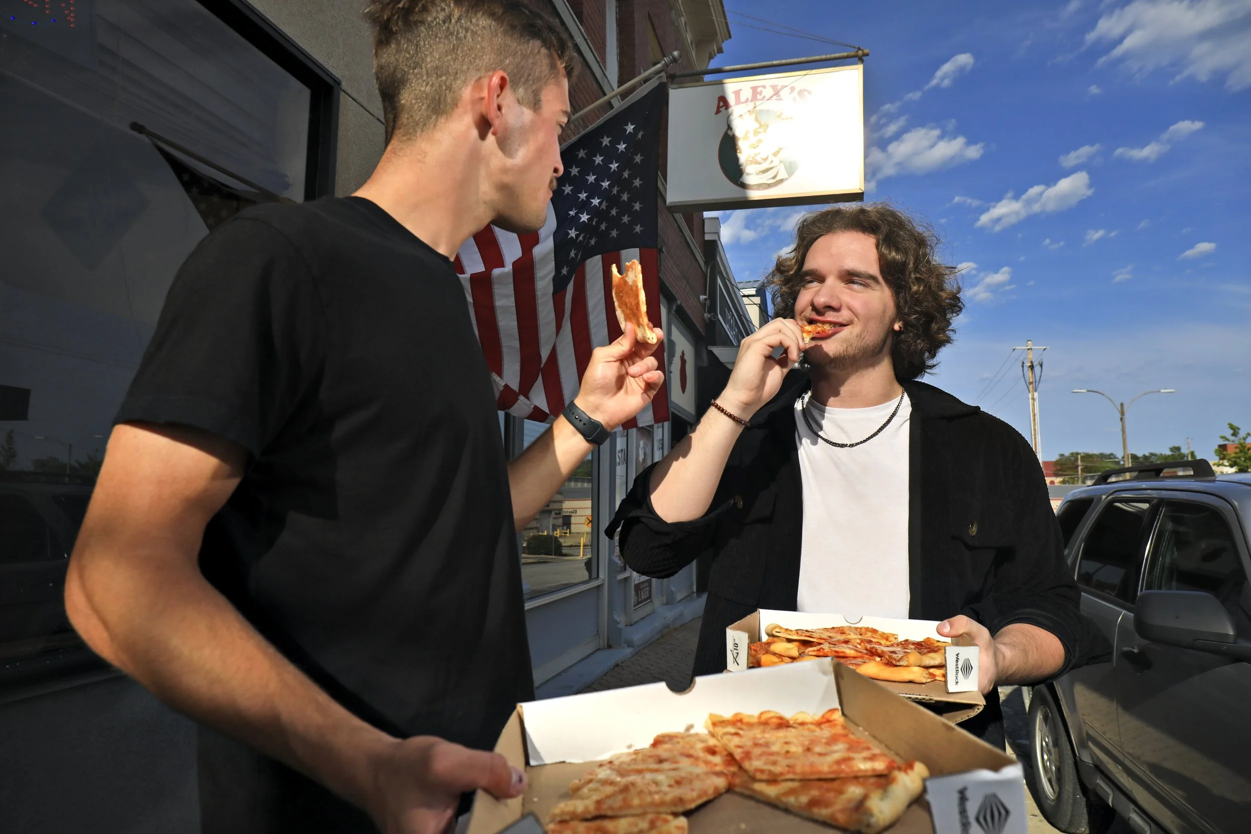 Two young men eating pizza outside a pizzeria on a sunny day. One is holding a box of pizza, and the other is holding a slice of pizza. They are standing near parked cars, with a building and a sign that says 'ALEX'S' in the background. An American f