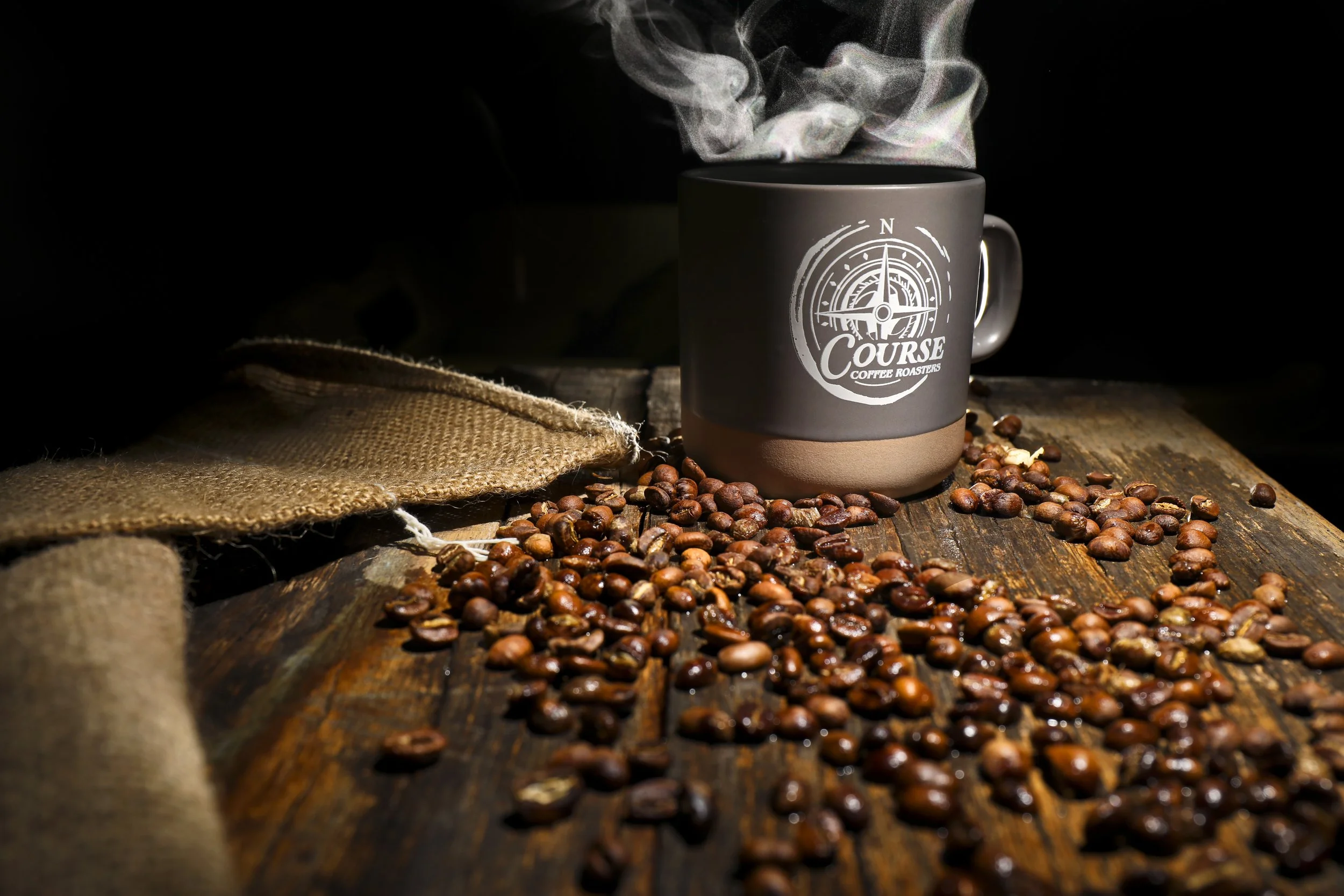 A steaming coffee cup with a logo, surrounded by scattered coffee beans on a rustic wooden table.