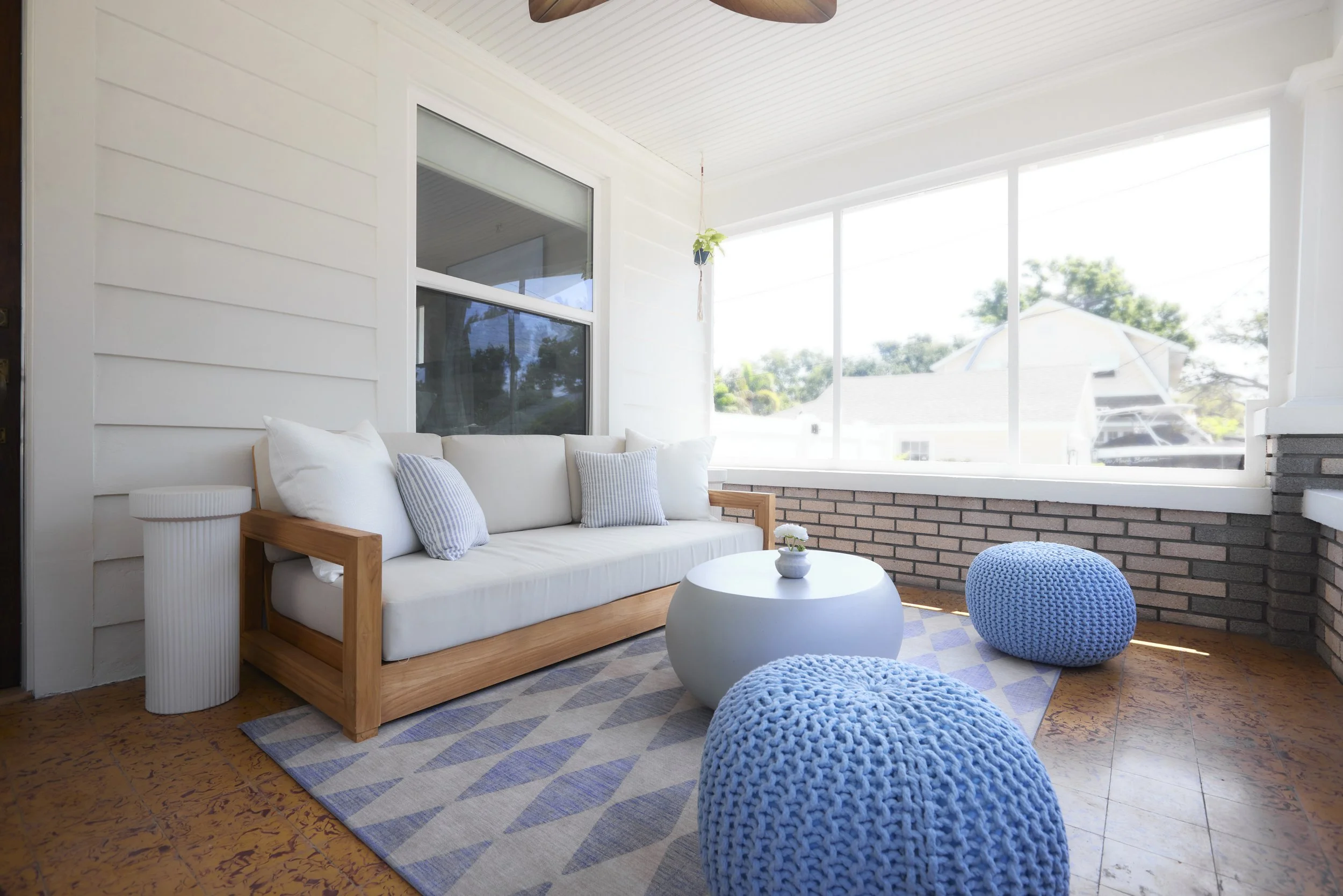 Sunroom with white wooden wall, cushioned wooden sofa, white coffee table, two knitted blue poufs, patterned rug, and large windows with outdoor view