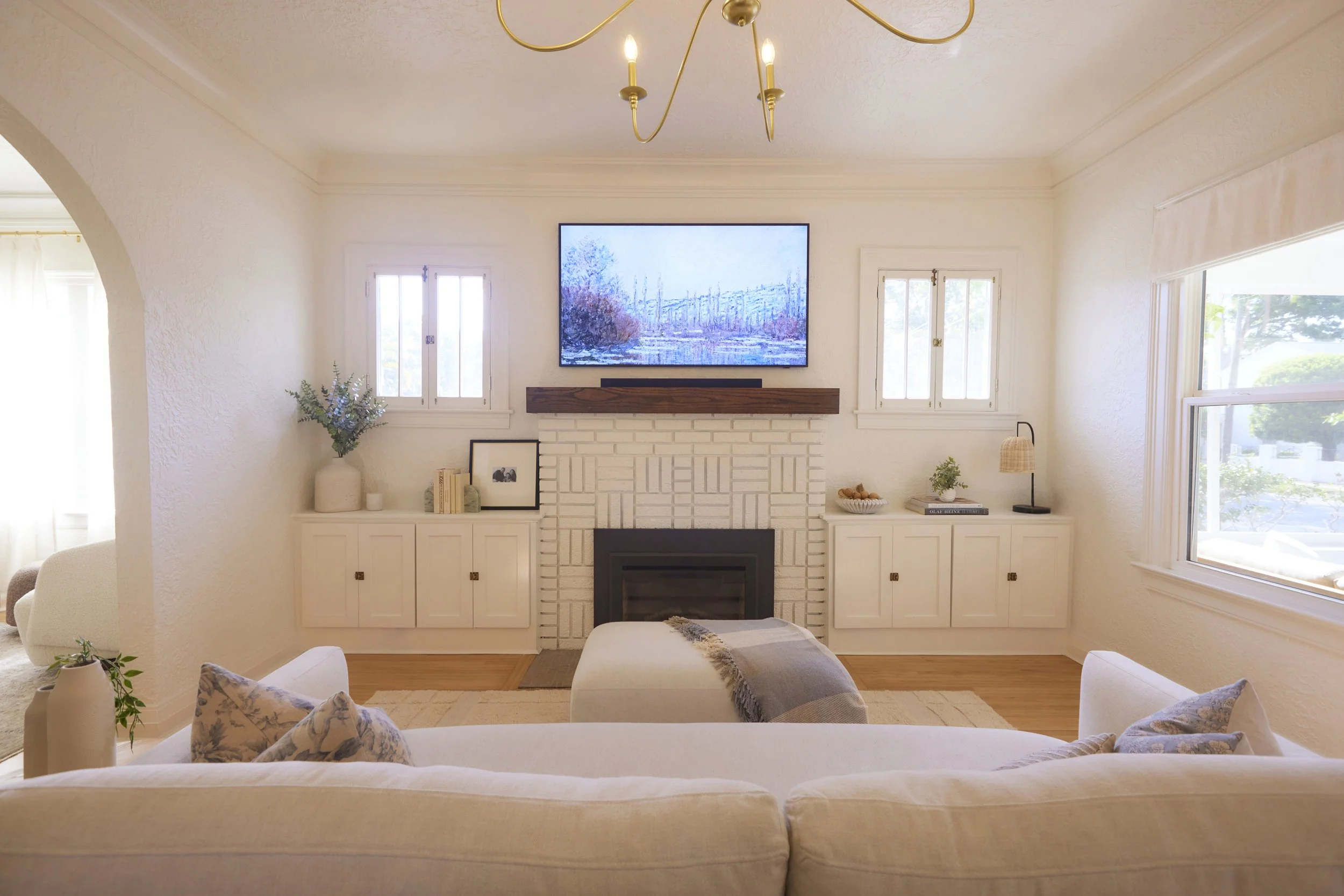Living room with white brick fireplace, TV mounted above, white built-in cabinets on each side, light beige sofa with decorative pillows, and large windows letting in natural light.
