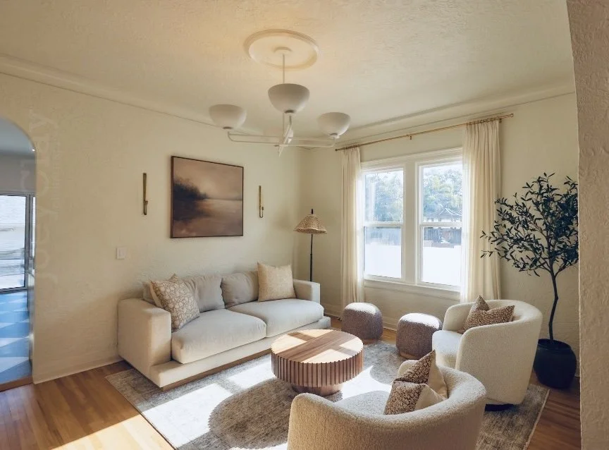 Bright living room with cream-colored sofa, two white armchairs, a round wooden coffee table, a standing lamp, a large window with white curtains, a framed landscape painting, a potted tree, and hardwood floors.
