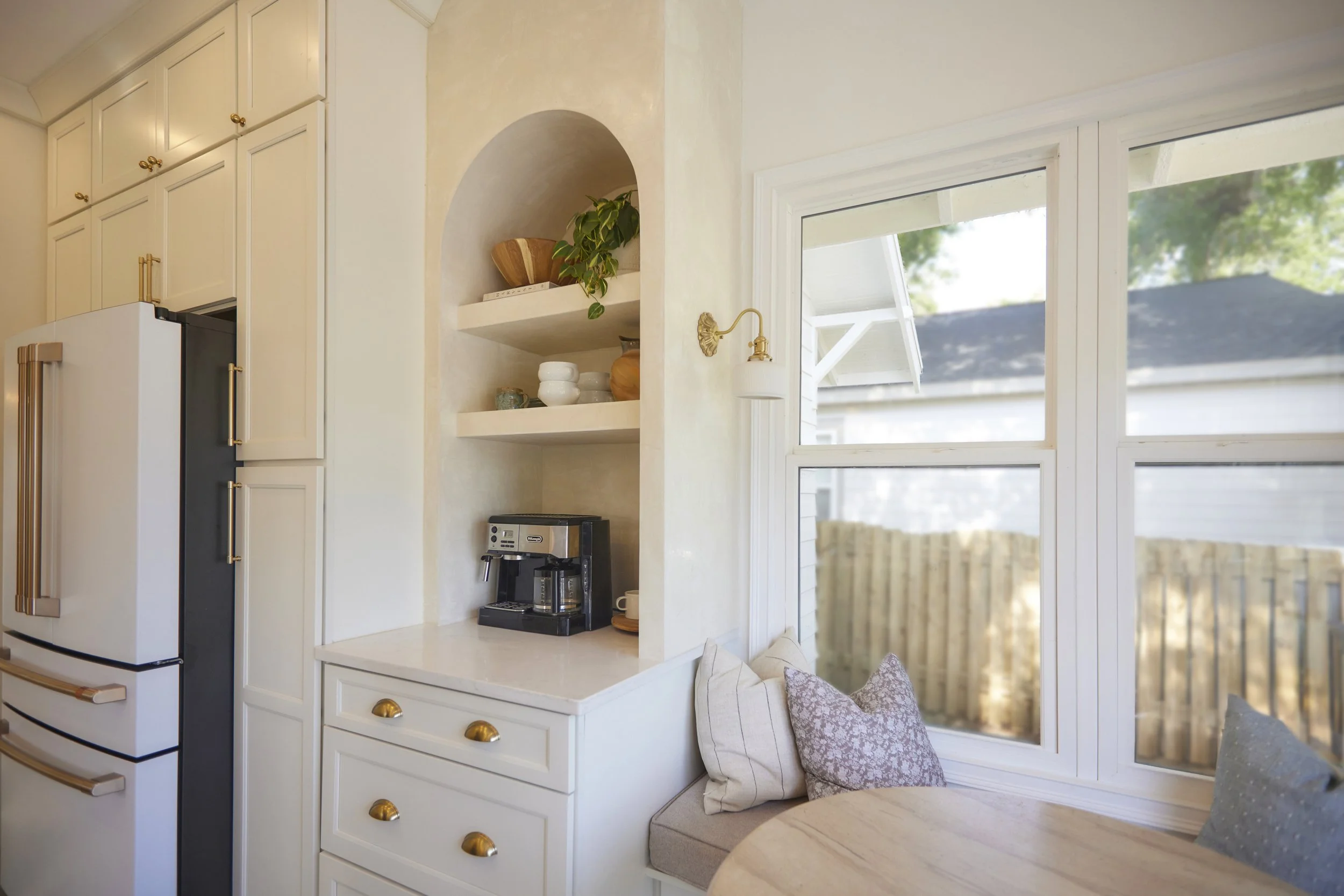 Kitchen corner with white cabinets, a coffee machine on the counter, a window seat with pillows, and large window showing an outdoor fence.