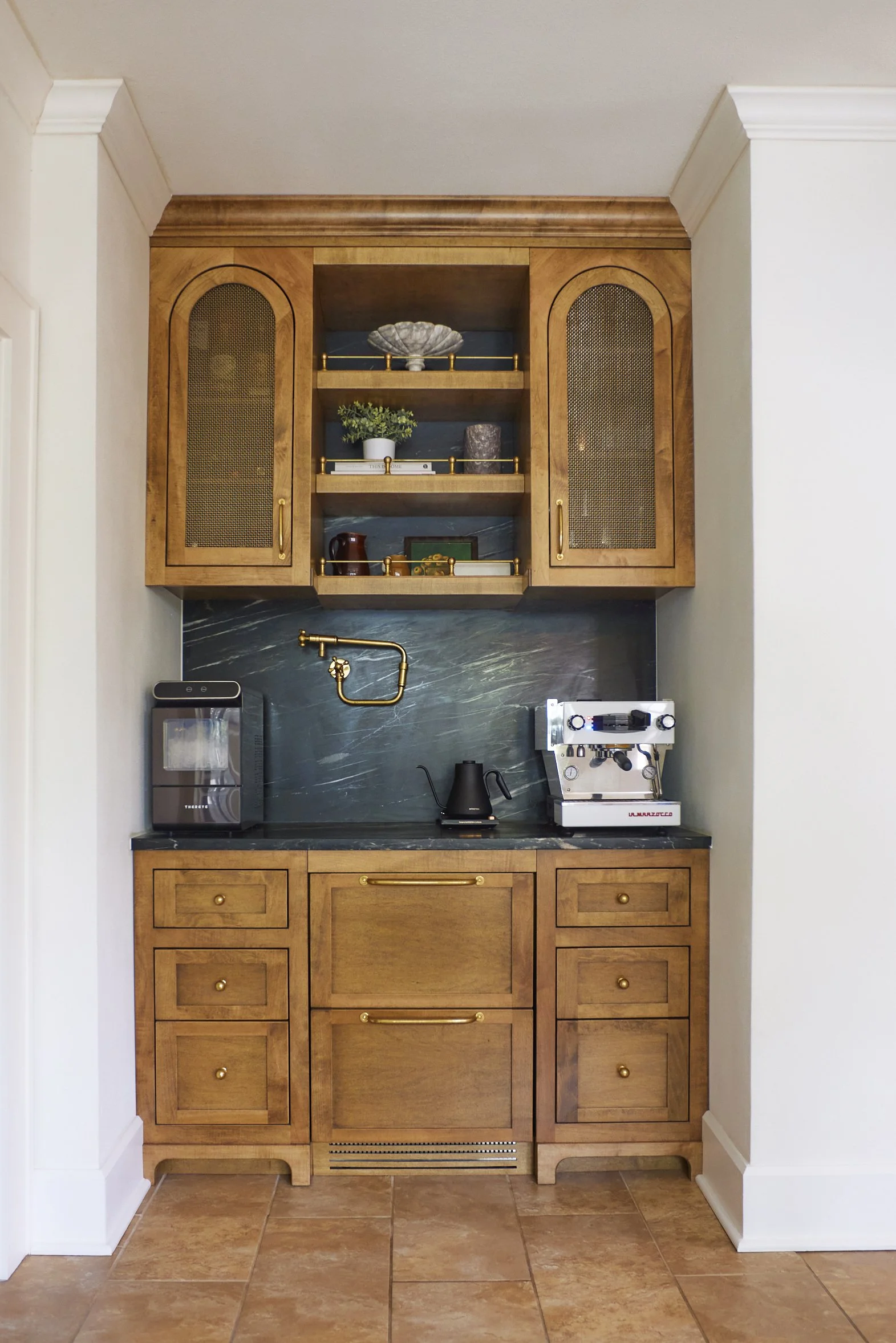 A small kitchenette with wooden cabinets, green marble countertop, and a backsplash. There is a coffee machine, a kettle, and a coffee grinder on the counter. Open shelving in the upper cabinet displays decorative items and books. A brass faucet is m