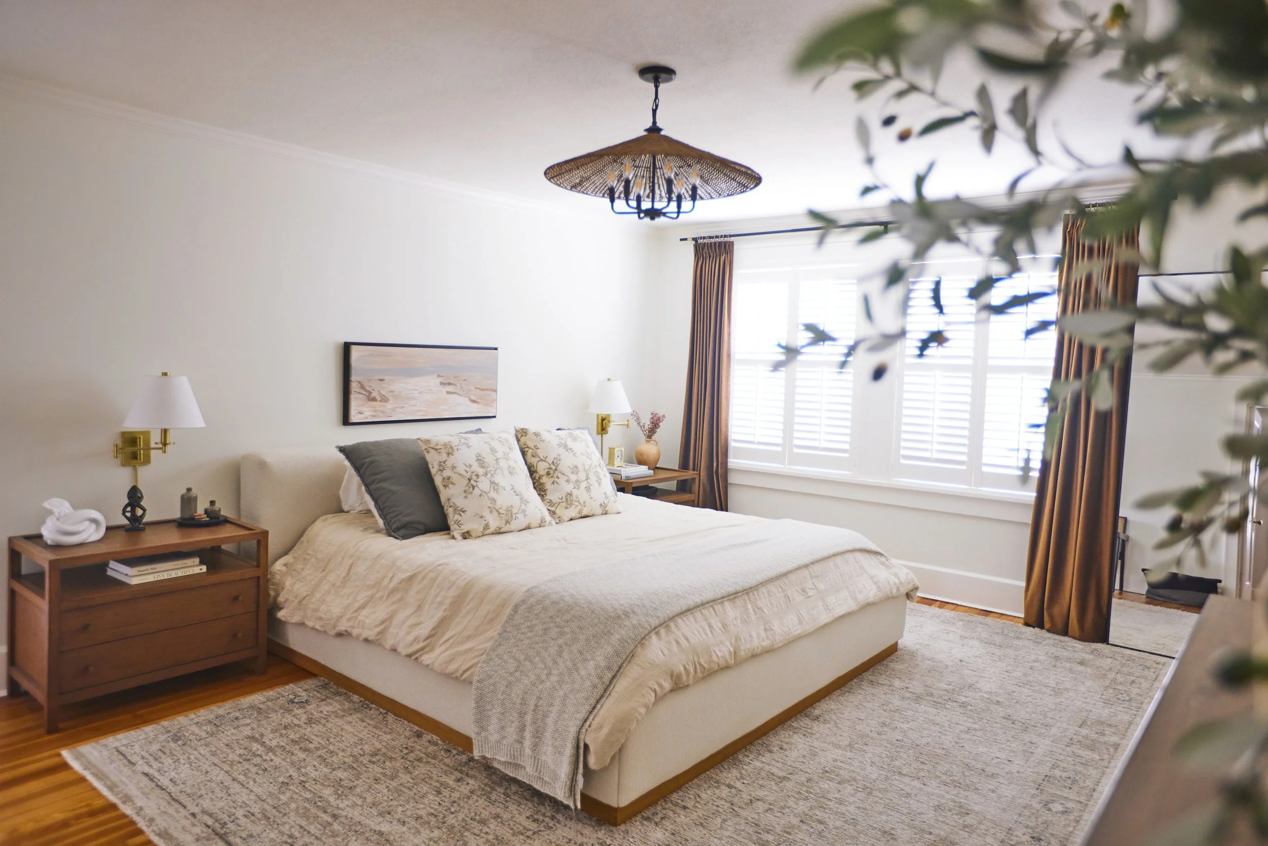 Bright bedroom with a bed, pillows, bedside tables, lamps, large window with blinds, and curtains.