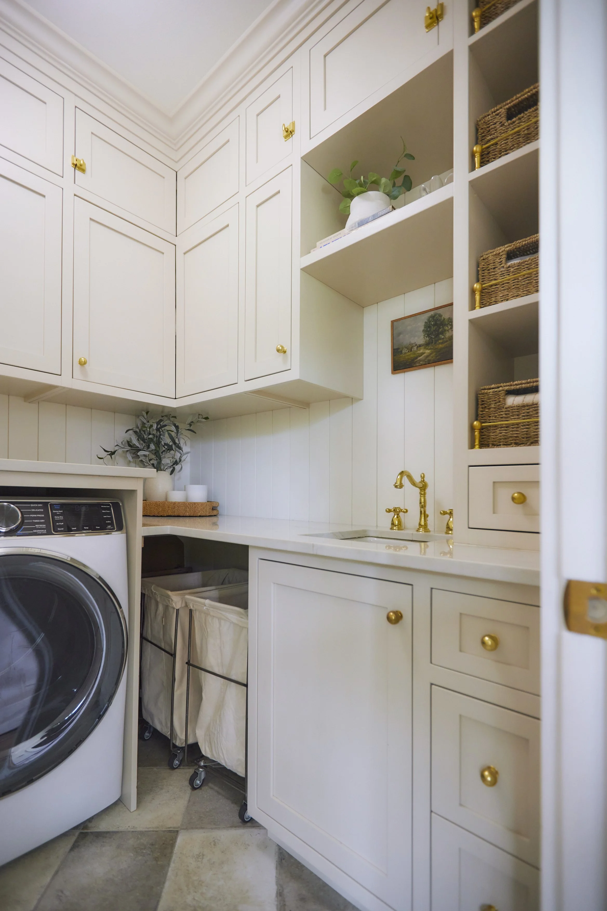 A laundry room with white cabinets, gold knobs, a small sink with gold fixtures, a washing machine, and wicker baskets on open shelves.