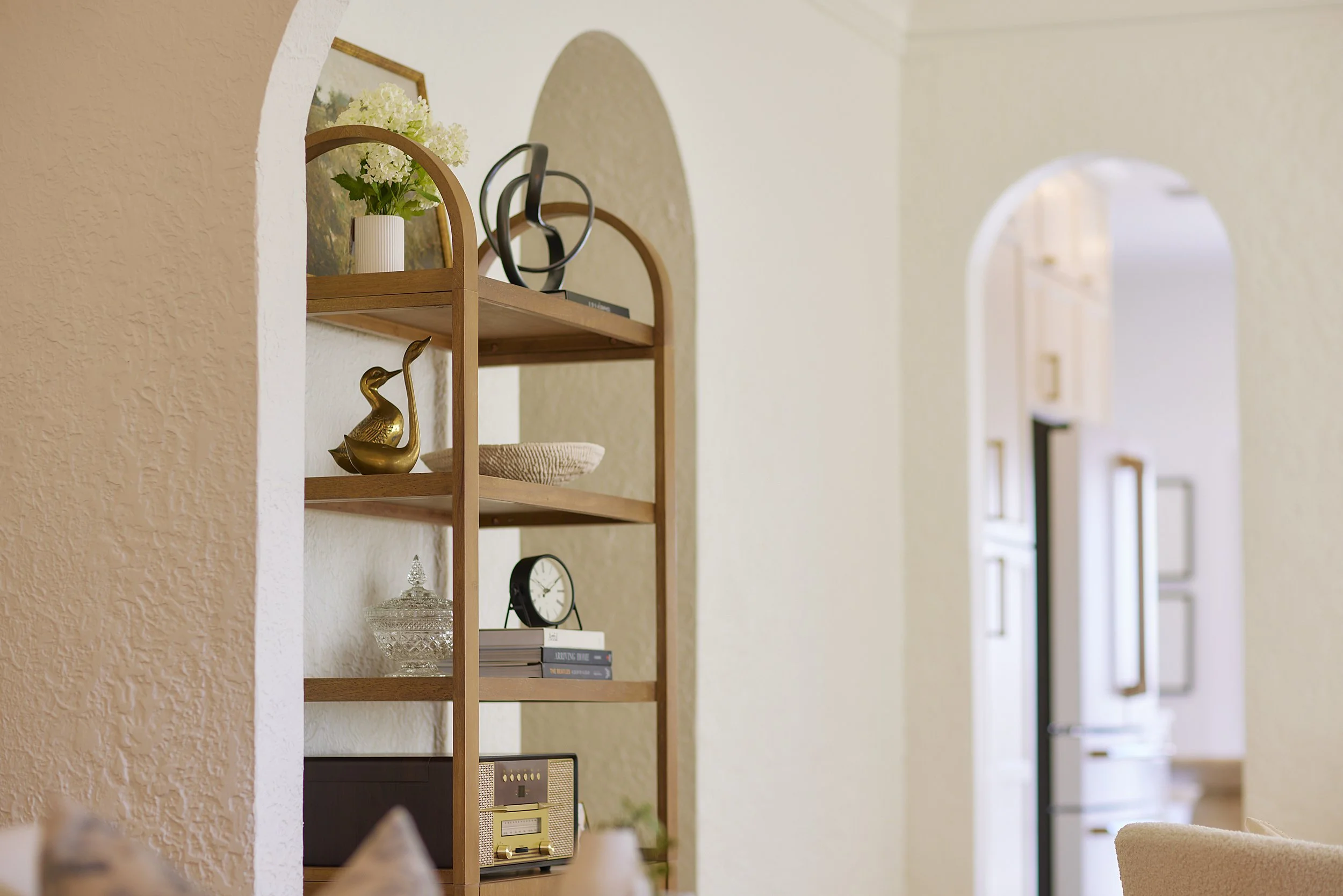 Living room with wooden bookshelf decorated with vases, books, clock, and decorative items, with an arched doorway to a kitchen area.