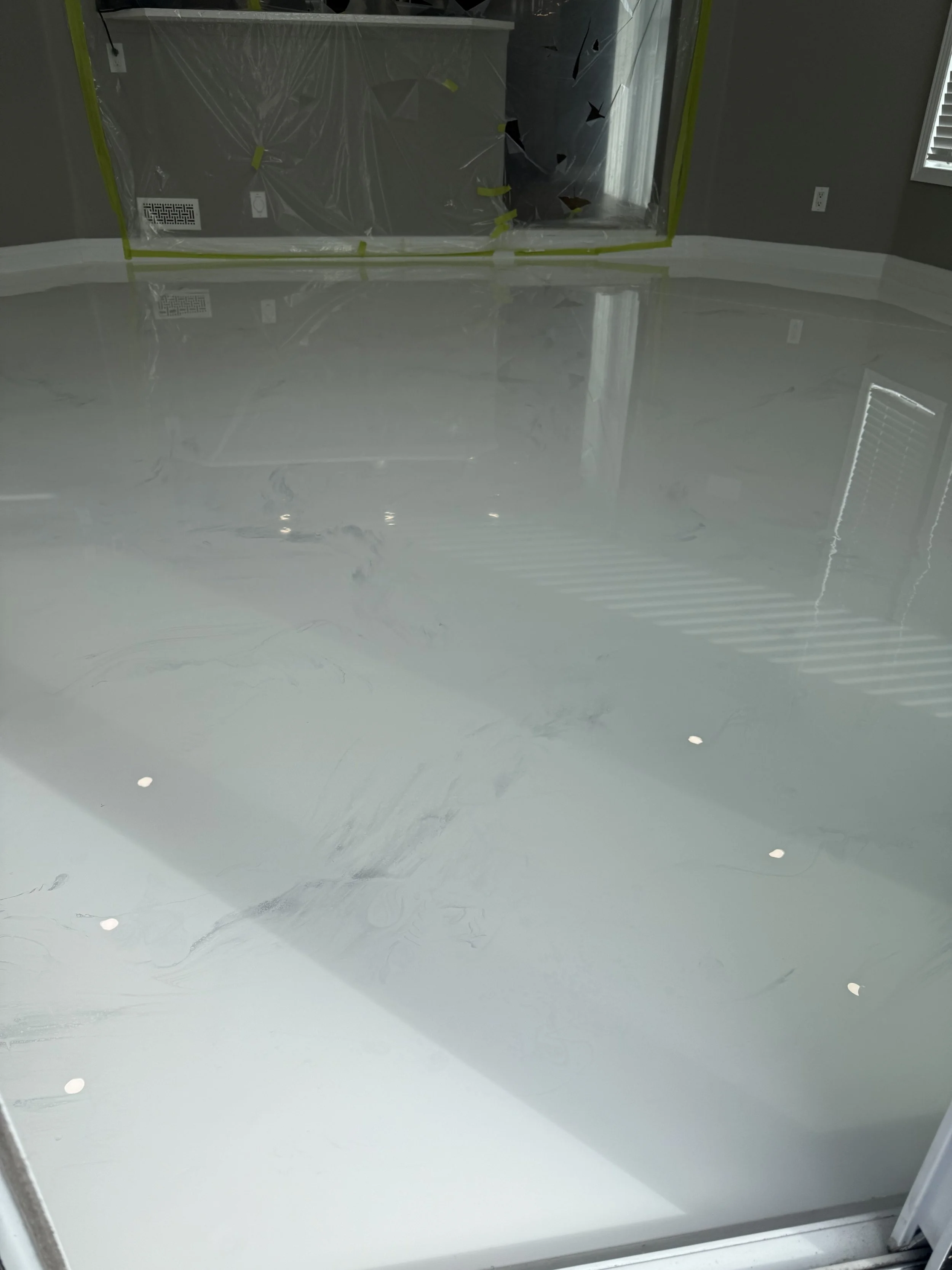 Freshly painted or coated light gray veins marble epoxy floor in a room under renovation, with protective plastic covering part of the wall and plastic on the floor, and a window with blinds.