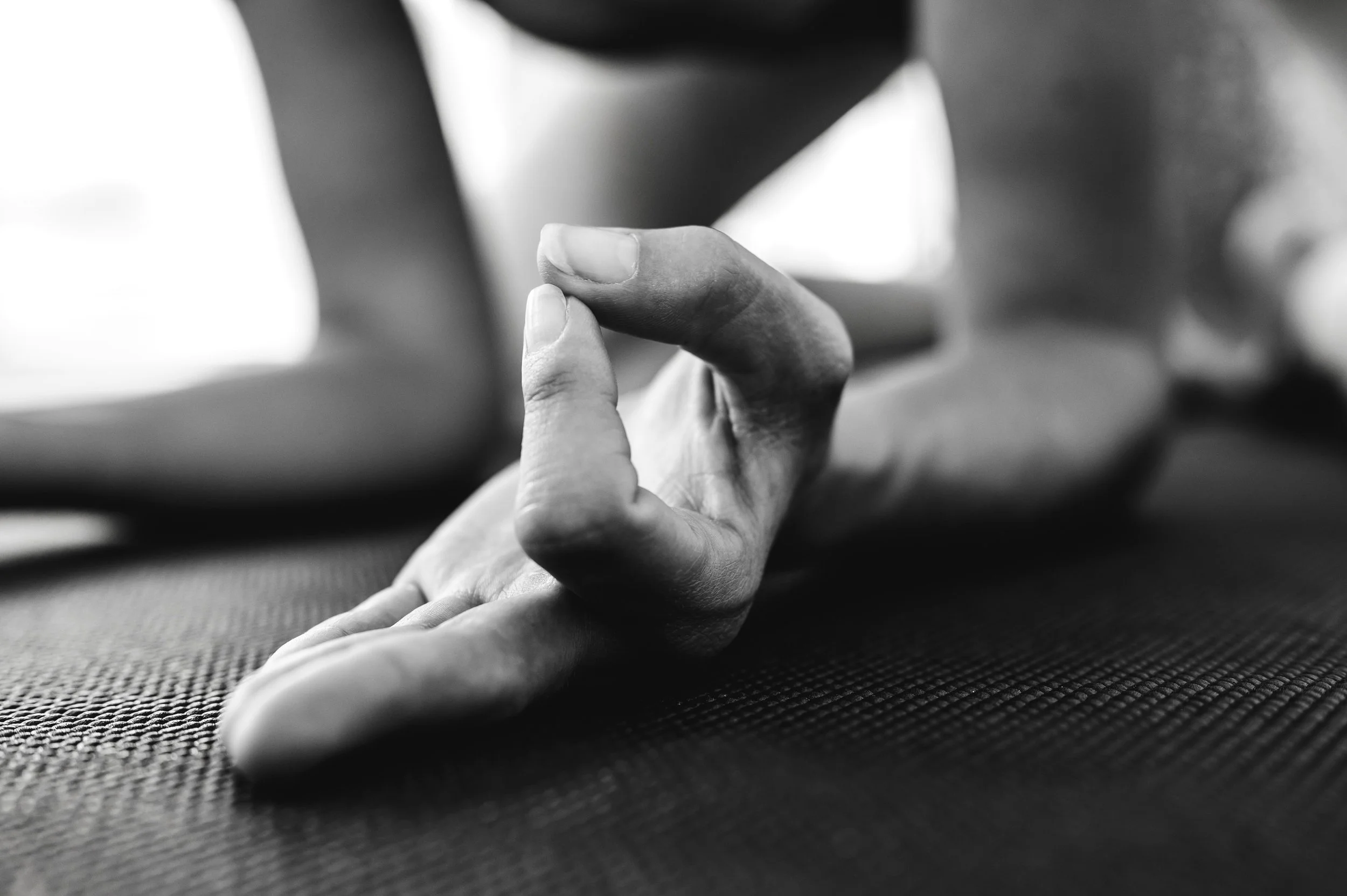 A close-up black and white photo of a person performing a yoga or meditation pose on a textured mat, with focus on their hand forming a mudra.