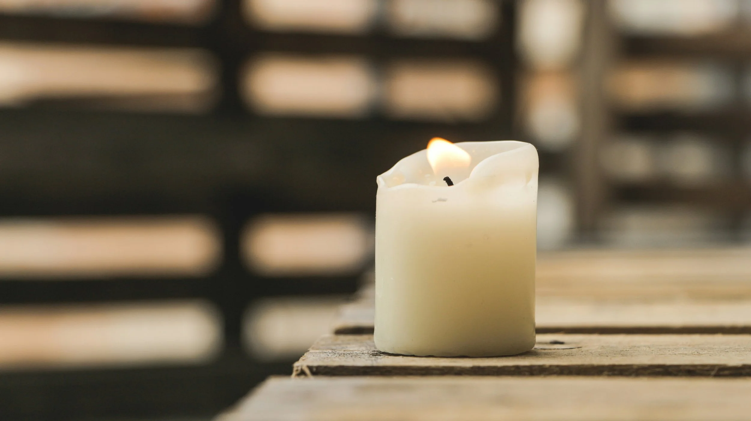 A lit white candle with a partially melted top sits on a wooden surface, with a blurred background.