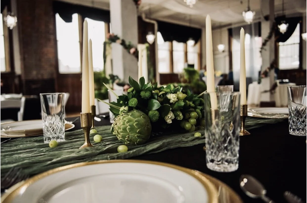 Elegant dining table with a green floral centerpiece, candles, crystal glasses, white plates with gold trim, set in a decorated venue with large windows and hanging lights.