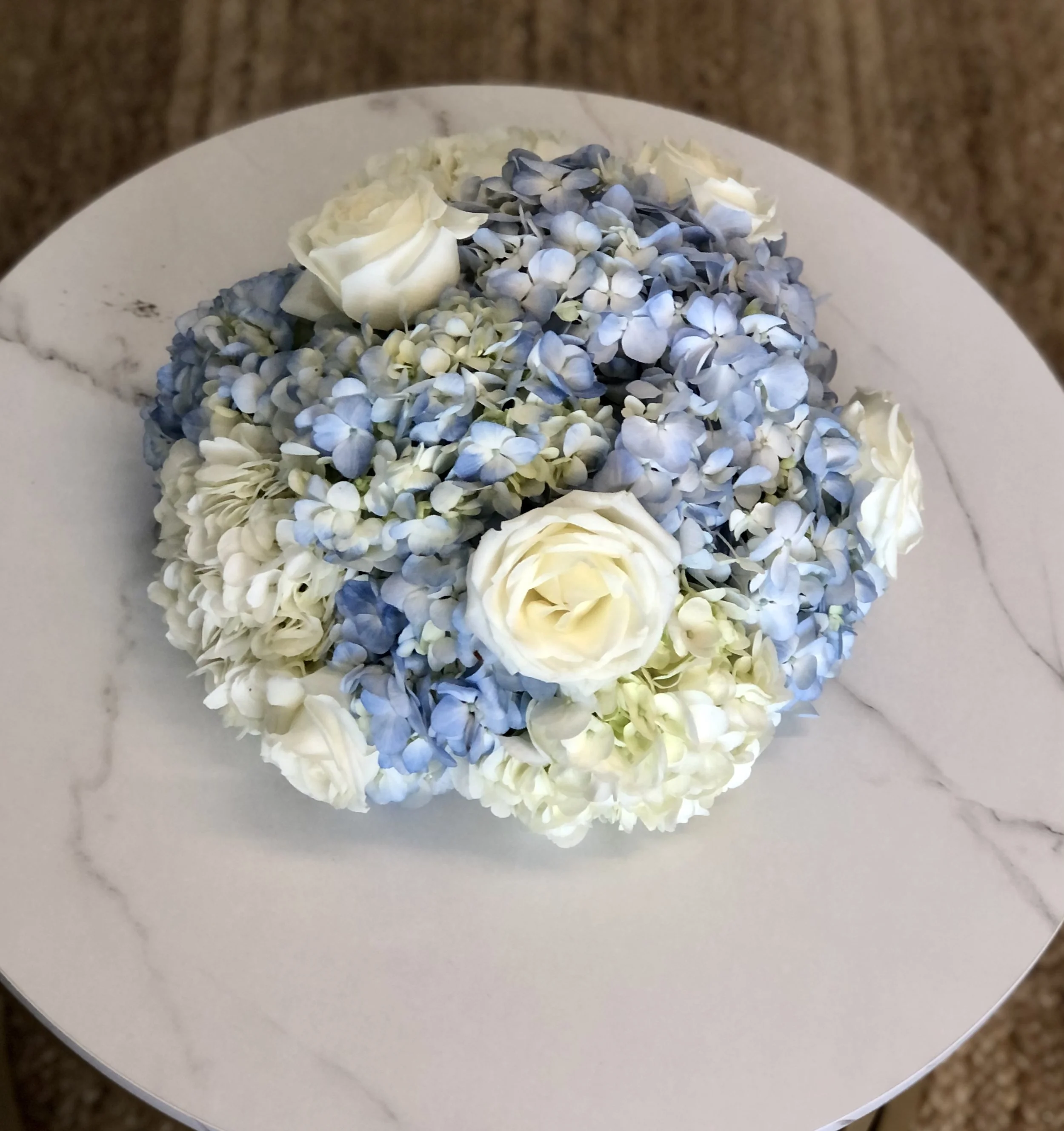 A bouquet of white roses, light blue hydrangeas, and white carnations on a white marble surface.