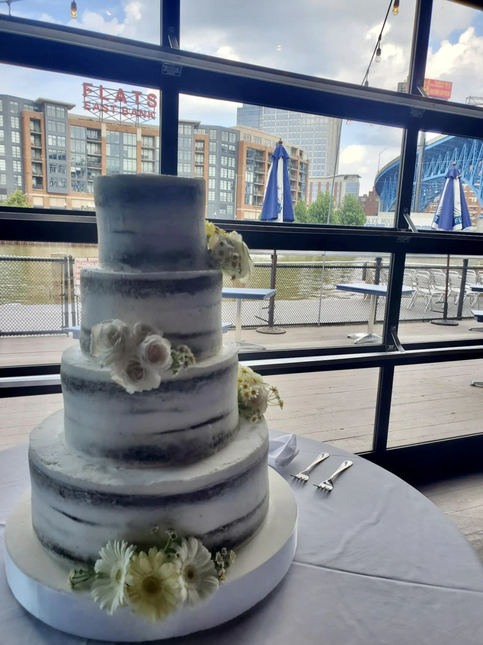 A four-tiered semi-naked wedding cake decorated with white and pink flowers, placed on a table with silver cake knives, in front of large windows overlooking a cityscape with buildings, umbrellas, and a bridge.