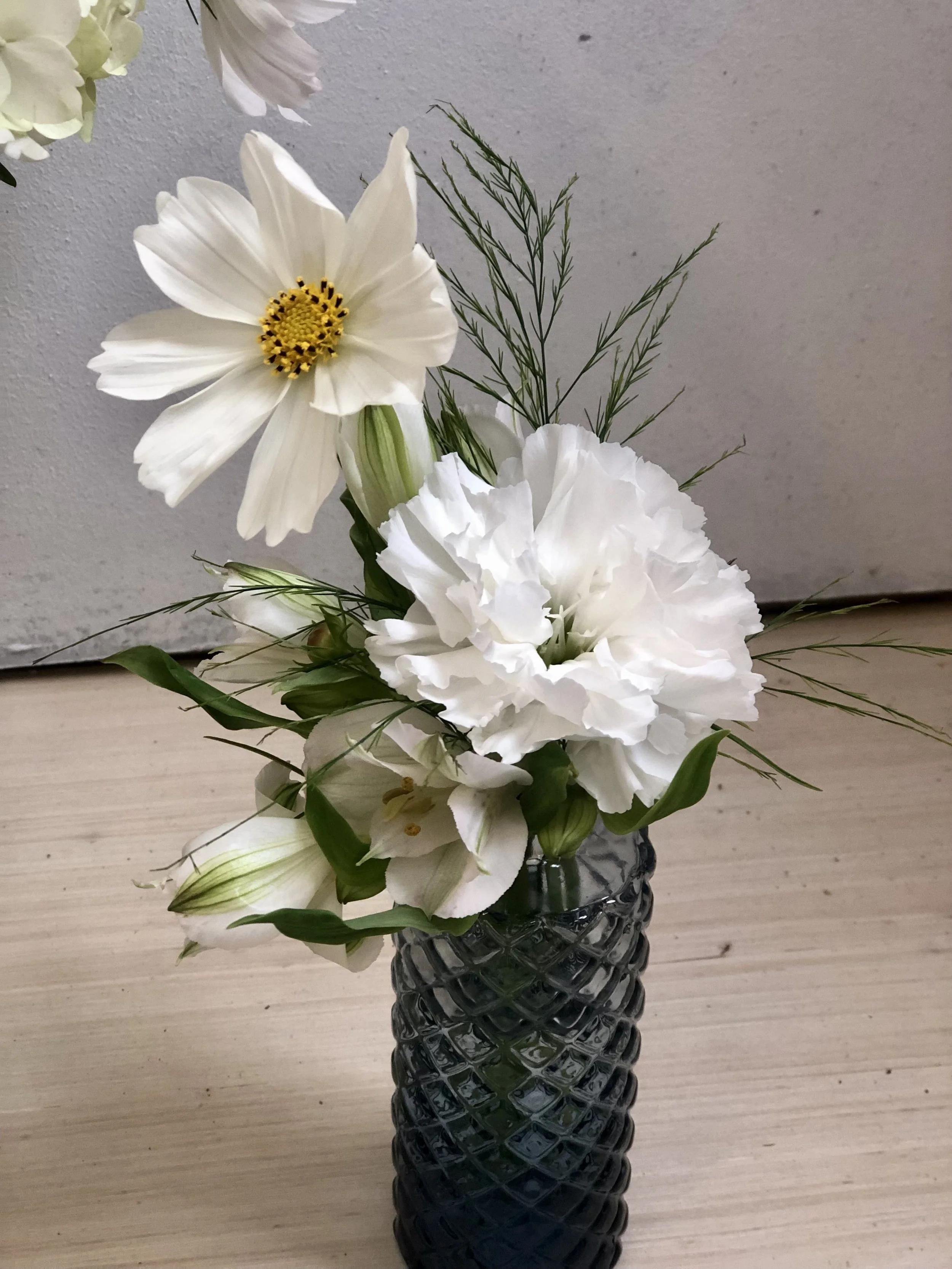 White flowers in a dark textured glass vase on a wooden surface against a light gray wall.