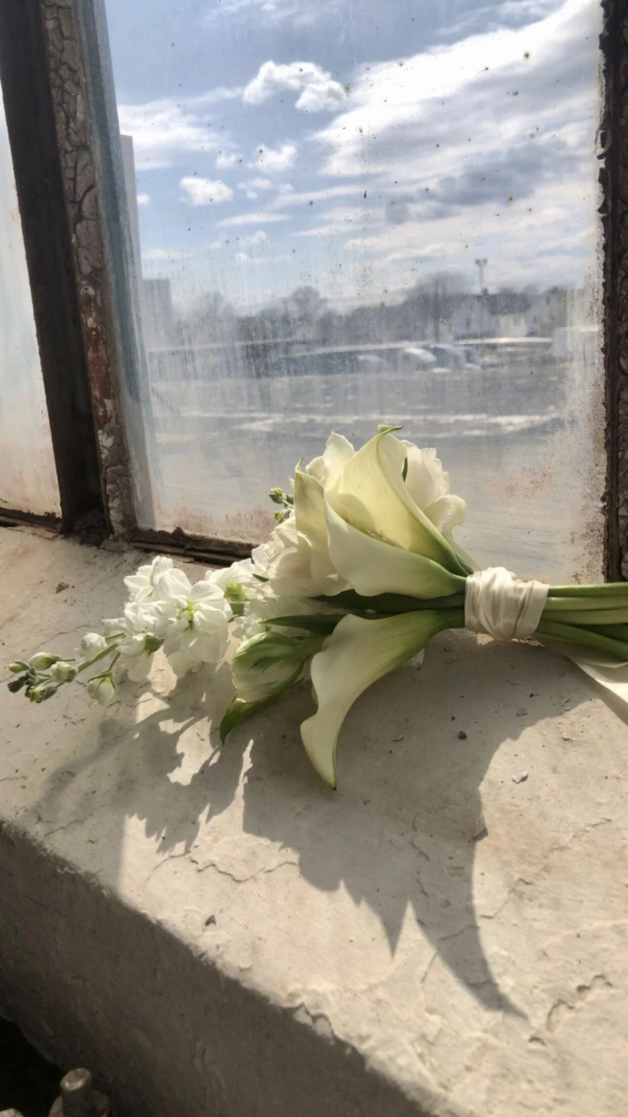 A white bouquet of flowers, including lilies and stock, resting on a windowsill with a view of a cloudy sky and parking lot outside.