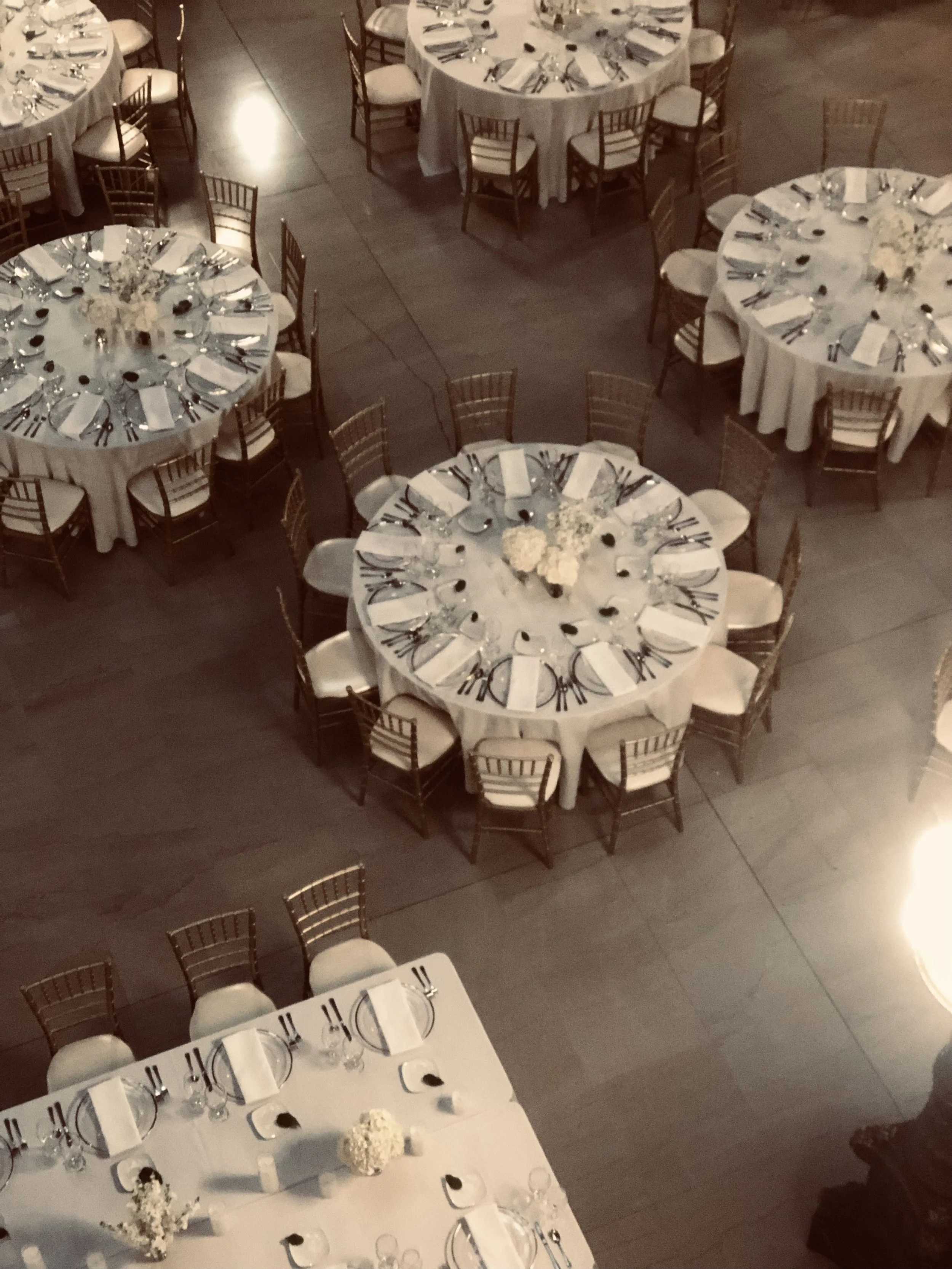 Elevated view of multiple round and rectangular banquet tables set for a formal event with white tablecloths, neatly arranged silverware, glasses, plates, and floral centerpieces, arranged on a wooden floor.