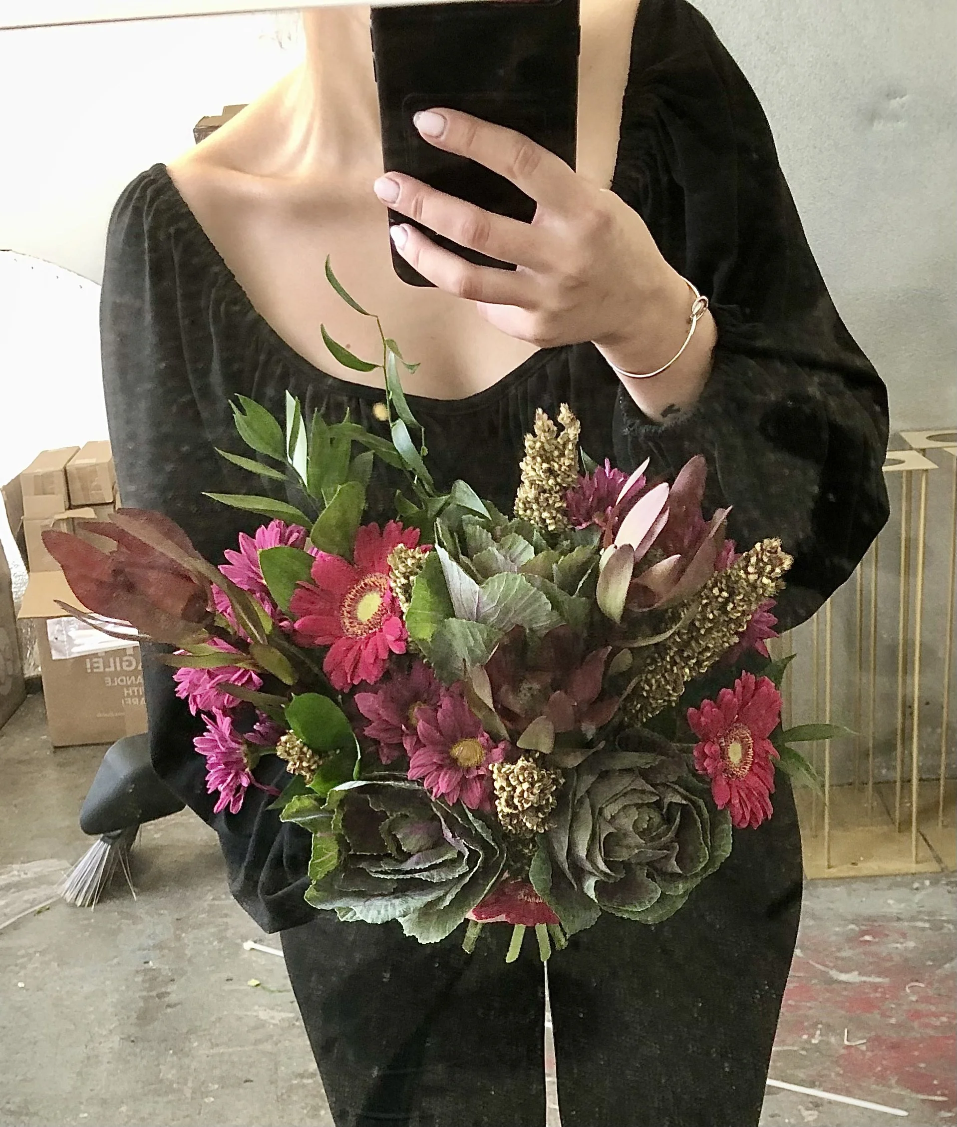 Person in black top taking a mirror selfie, holding a bouquet of pink, green, and purple flowers.