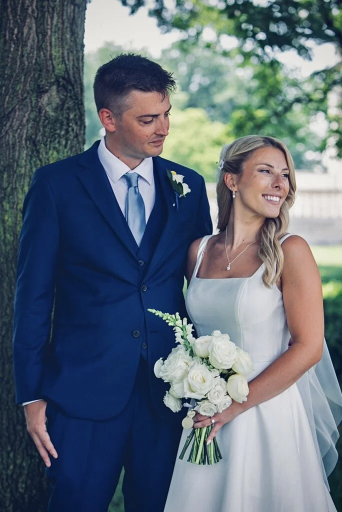 A bride and groom stand outdoors near a tree. The groom wears a navy blue suit with a light blue tie, and the bride wears a white wedding dress and holds a bouquet of white roses. The bride is smiling and looking slightly away, while the groom looks
