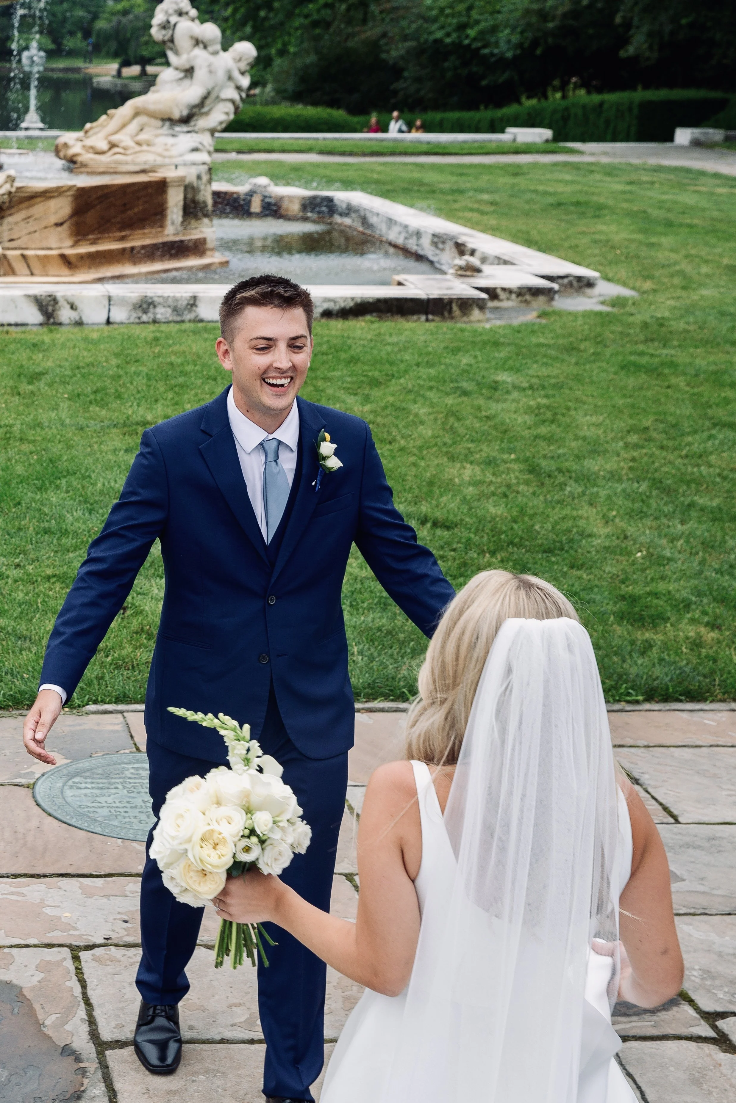 A groom in a dark blue suit and a bride with a white dress and veil sharing a joyful moment outdoors near a fountain and garden, with the bride holding a bouquet of white flowers.