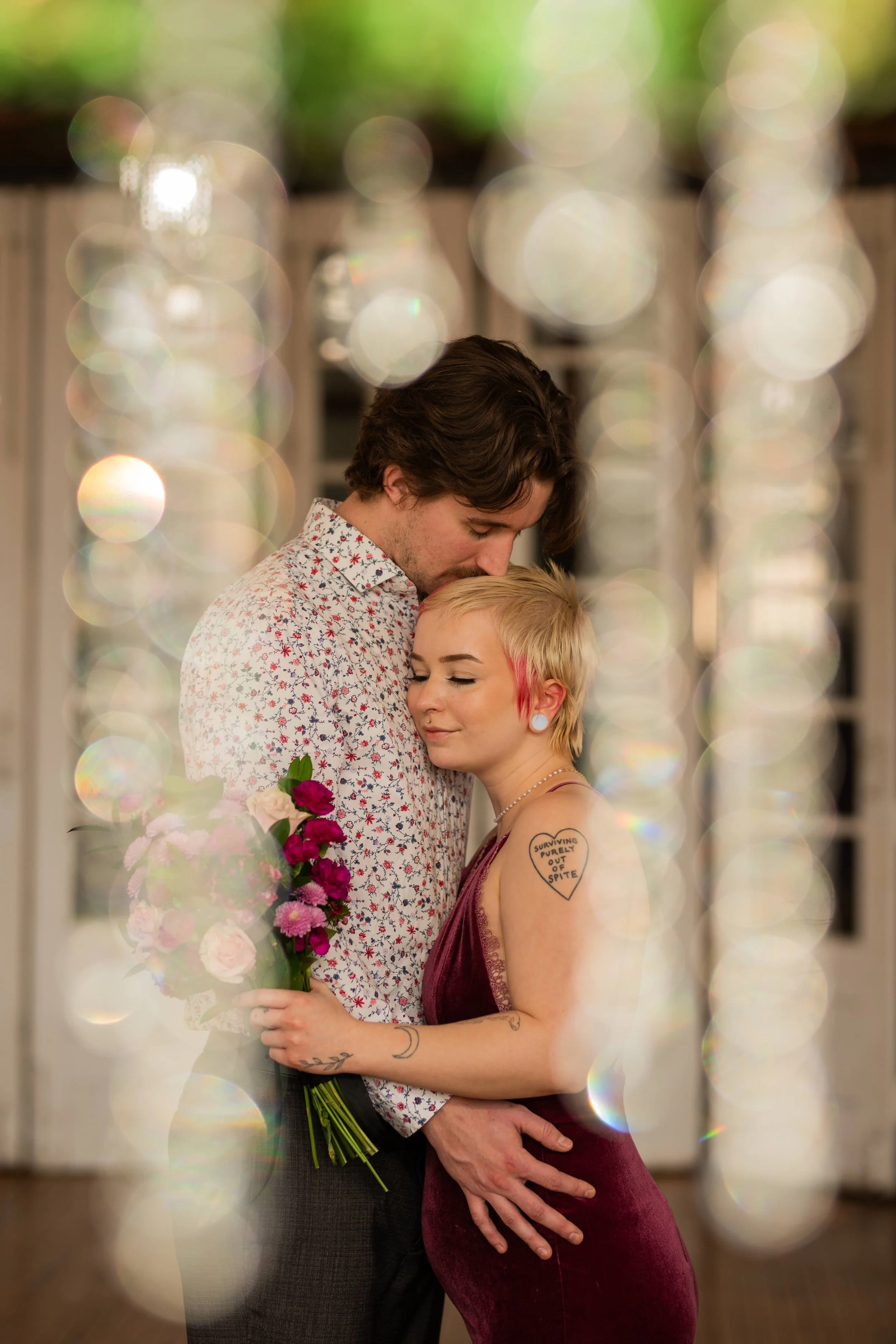 A couple embracing, with the woman holding a bouquet of flowers, and the man resting his forehead against her hair, framed by blurred light reflections.