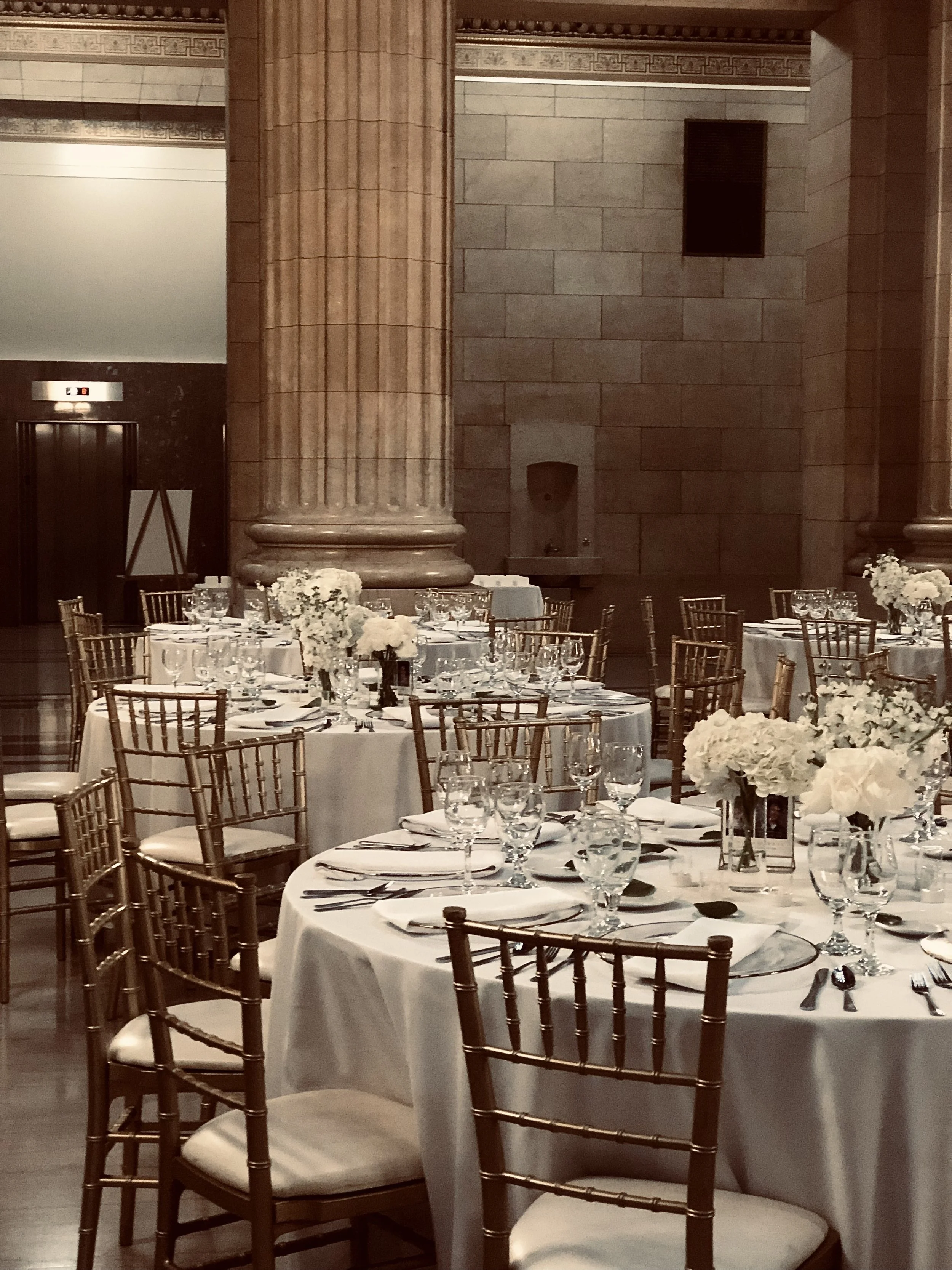Elegant event space with round tables covered in white tablecloths, decorated with floral centerpieces, and set with glasses, plates, and silverware. Gold Chiavari chairs surround the tables, and a large stone pillar is prominent in the background.