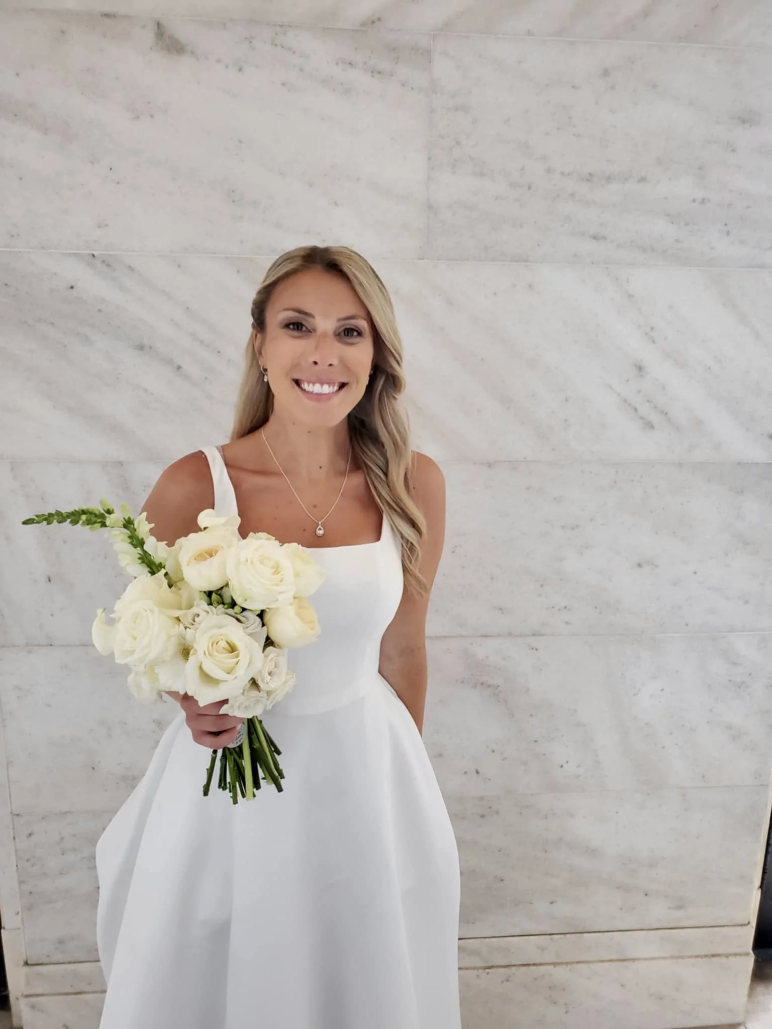 A smiling woman in a white wedding dress holding a bouquet of white roses, standing against a light-colored stone wall.