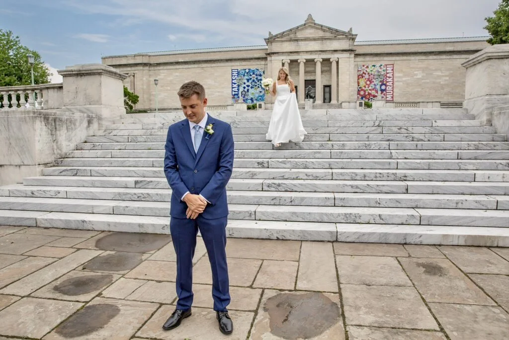 A bride and groom at a wedding in front of a historic building with wide marble stairs. The groom in a blue suit stands at the bottom of the stairs looking down, while the bride in a white gown stands on the stairs holding a bouquet.