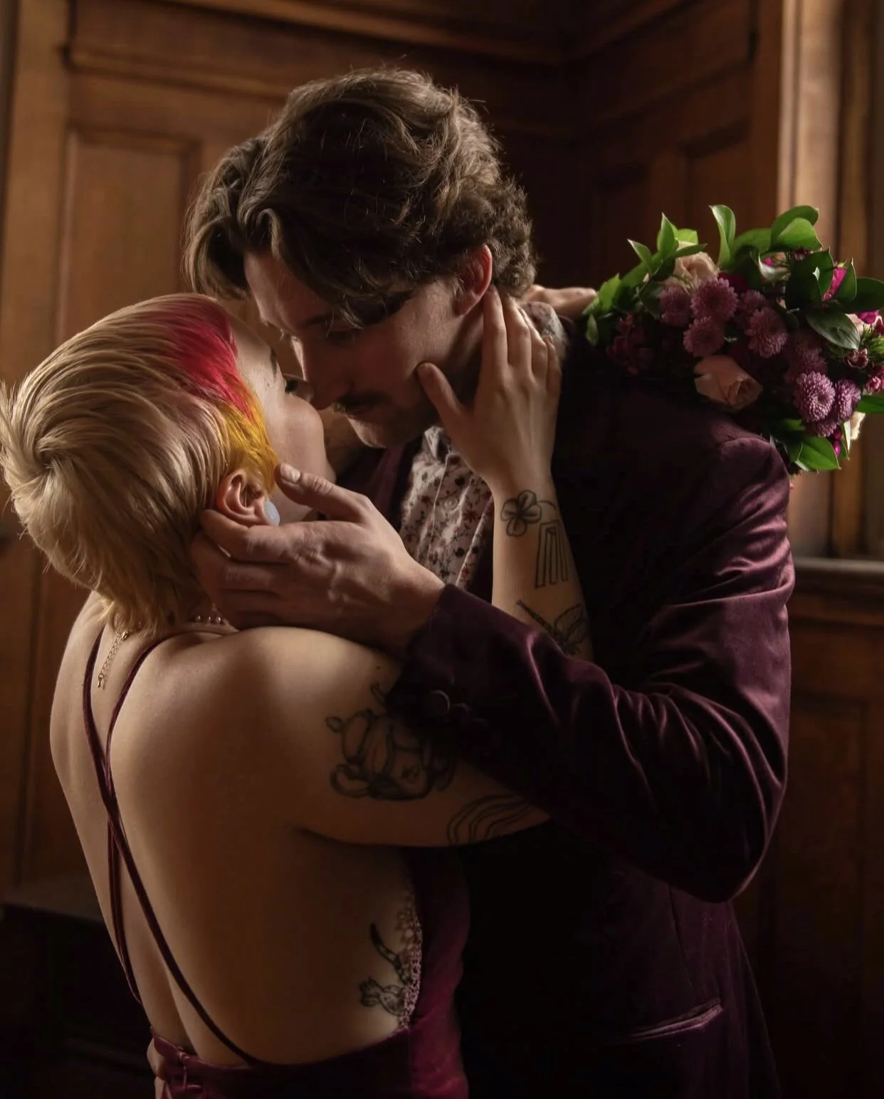A couple sharing a romantic kiss, the woman has colorful hair and tattoos, and is holding a bouquet of flowers.