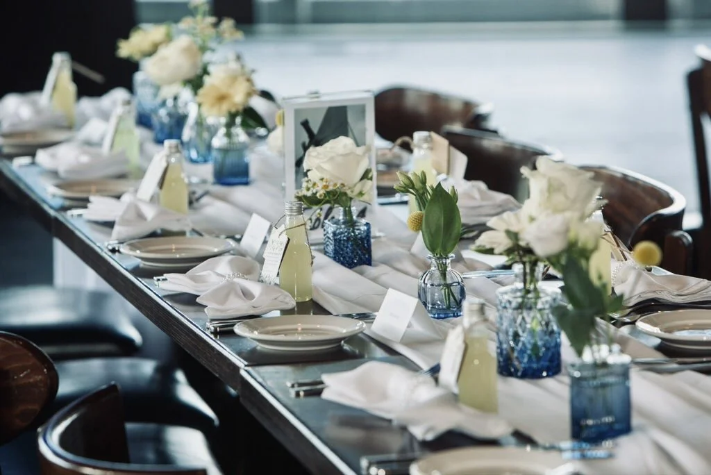 Elegant dining table set with white napkins, plates, floral centerpieces in blue vases, and bottles of pale yellow drink in glass bottles, with a white table runner.
