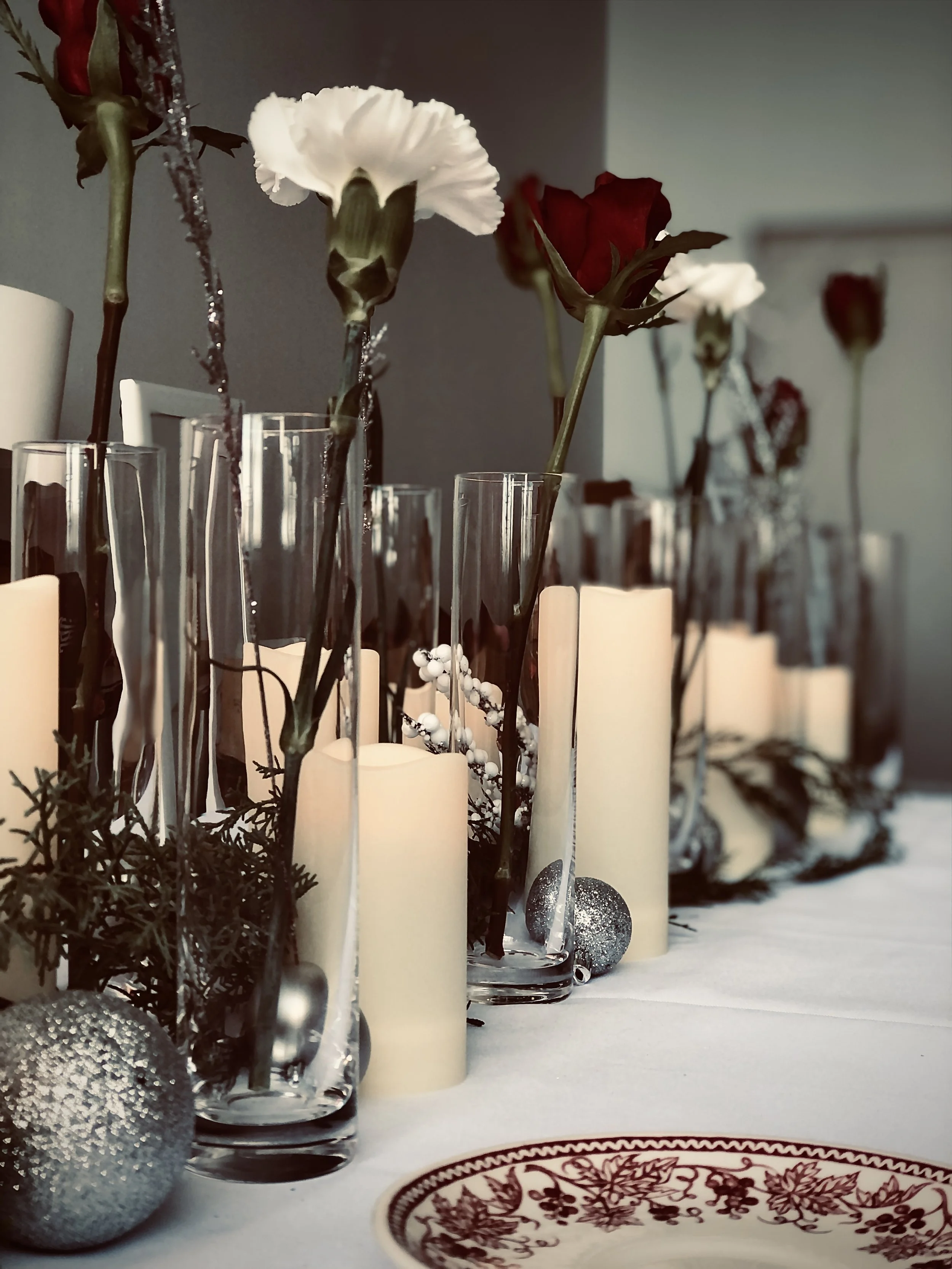Table decorated with white and red roses in glass vases, white candles, silver ornaments, and greenery, suggesting a festive or holiday setting.