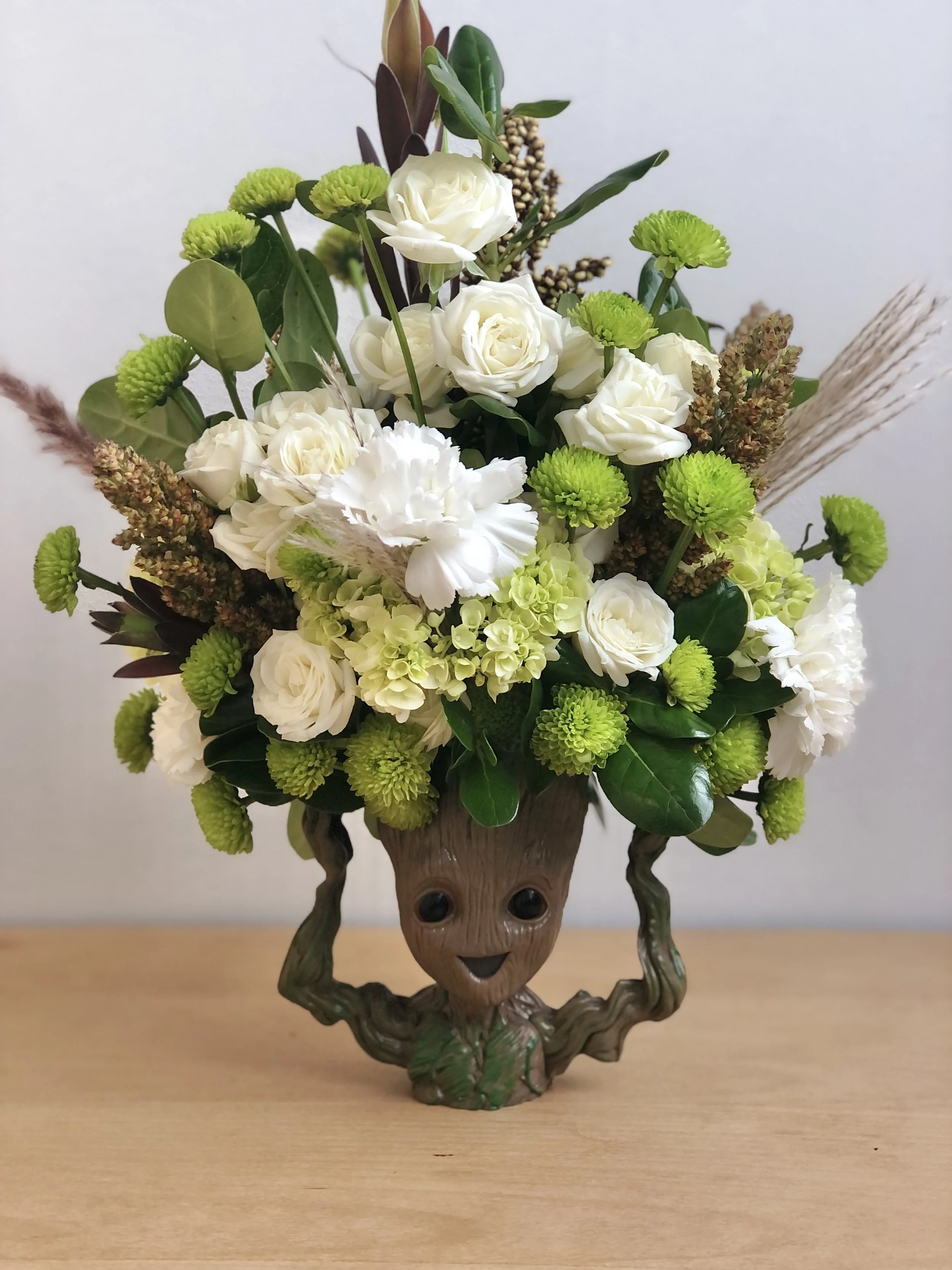 A decorative flower arrangement in a Groot-themed vase with white roses, green button poms, white carnations, hydrangeas, and greenery, placed on a wooden surface.