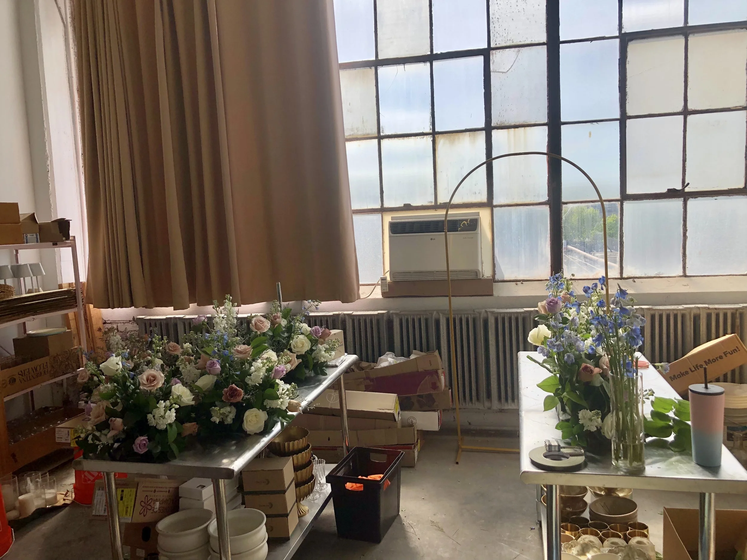 Two metal tables with floral arrangements in vases, surrounded by boxes, supplies, and a large window in an industrial or storage room setting.