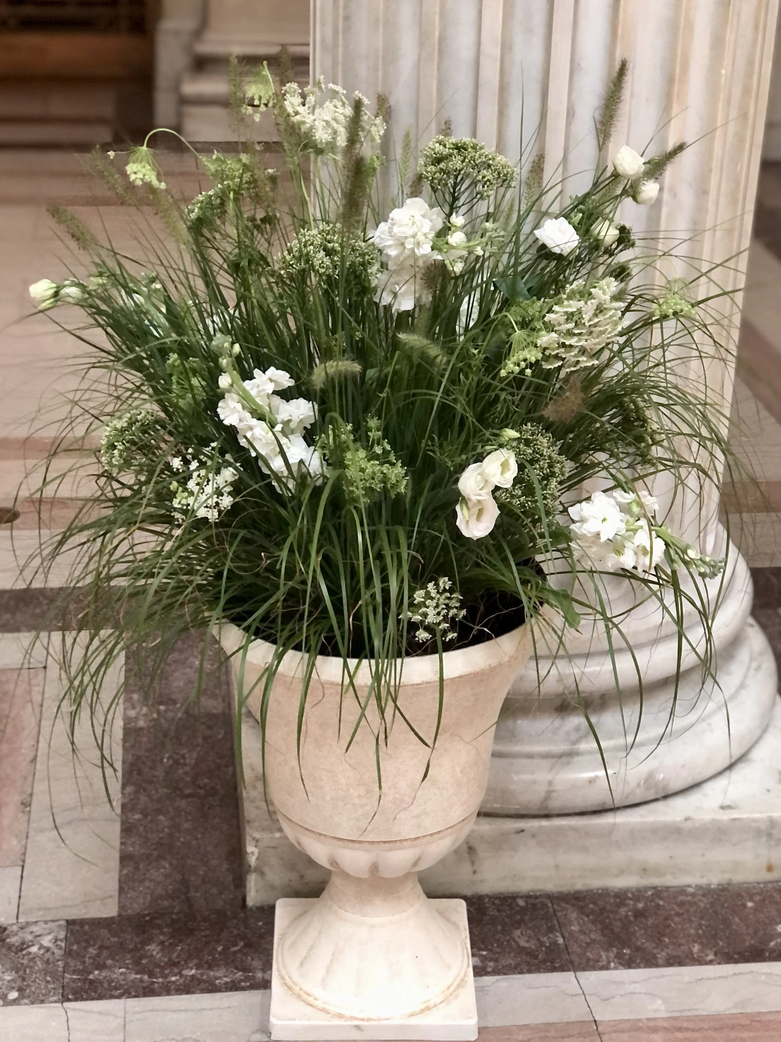 A large flower arrangement in a classical pedestal urn with white flowers and green foliage.