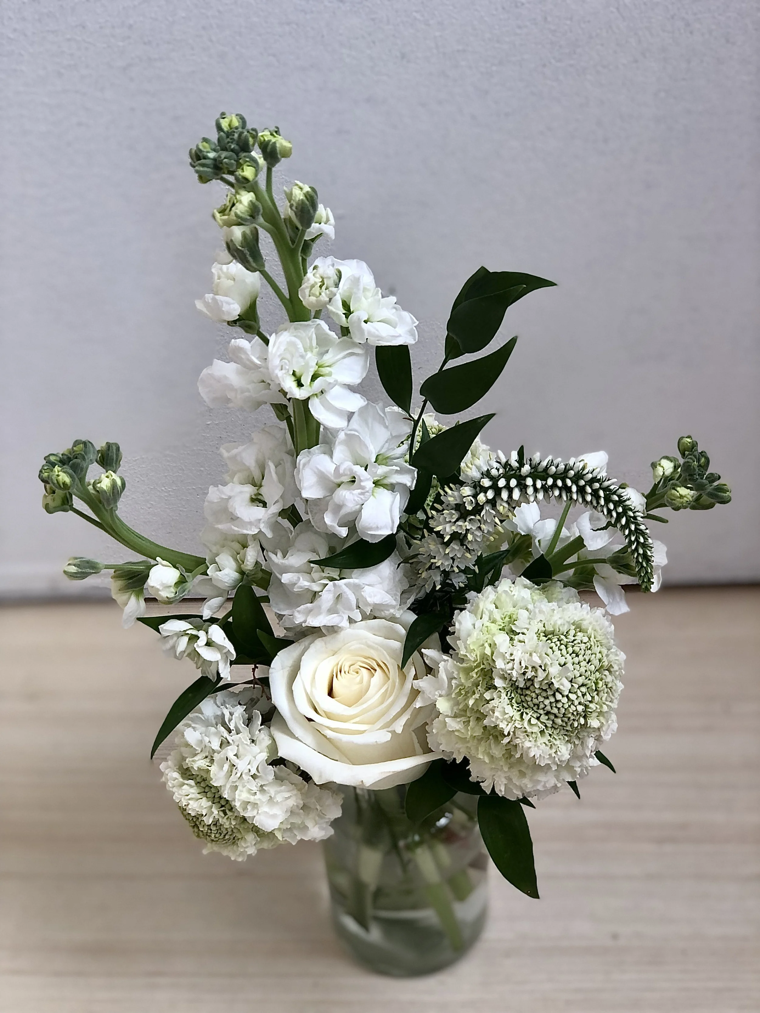 A bouquet of white flowers, including roses, carnations, and other blossoms, arranged in a clear glass vase with green leaves.