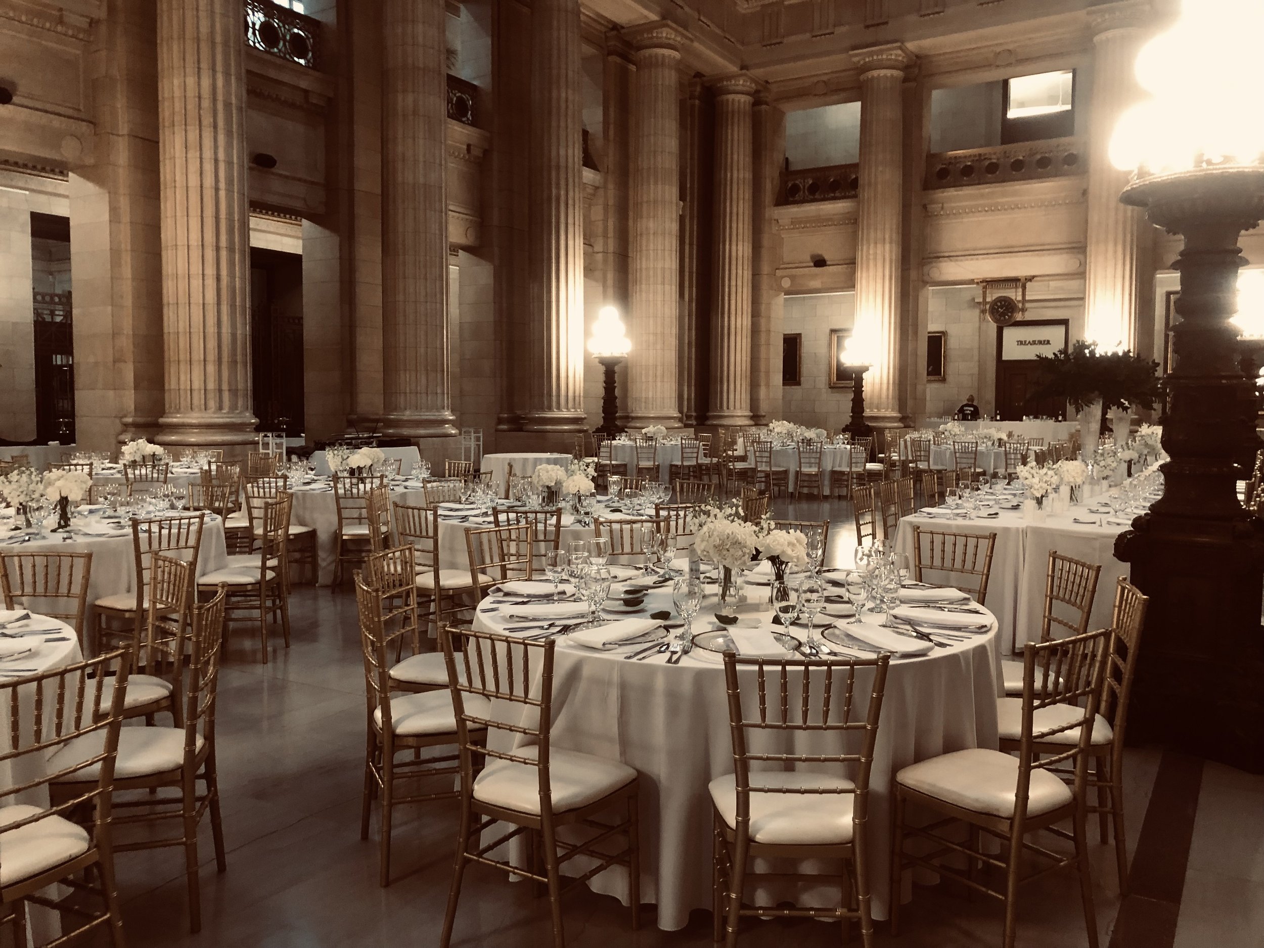 Elegant banquet hall with round tables covered in white tablecloths, decorated with white floral centerpieces, set with plates, glasses, and silverware, surrounded by gold chairs, in a grand room with tall columns and warm lighting.