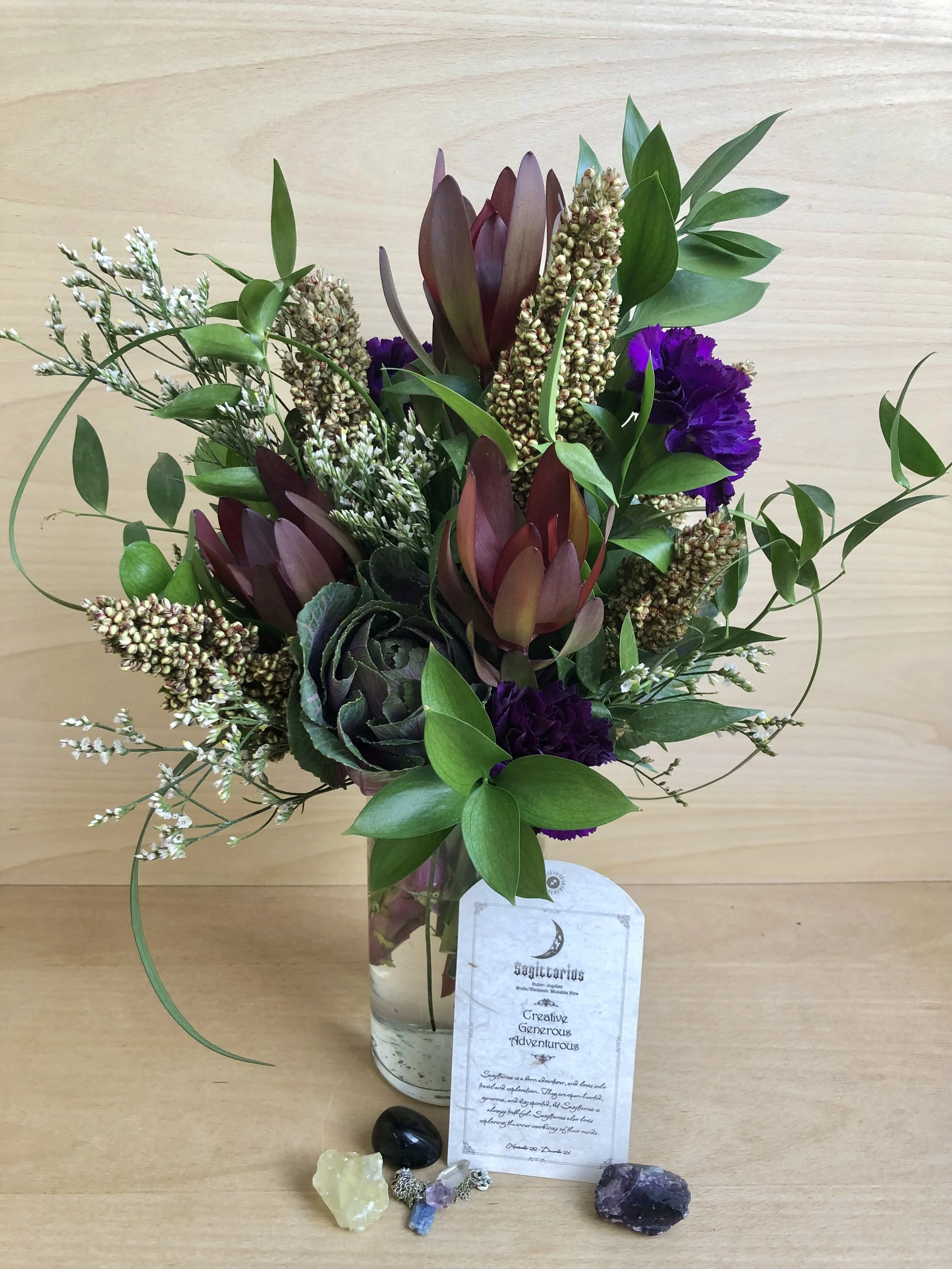 A bouquet of flowers in a glass vase with purple, green, and white blooms, along with attached crystals and a tag.