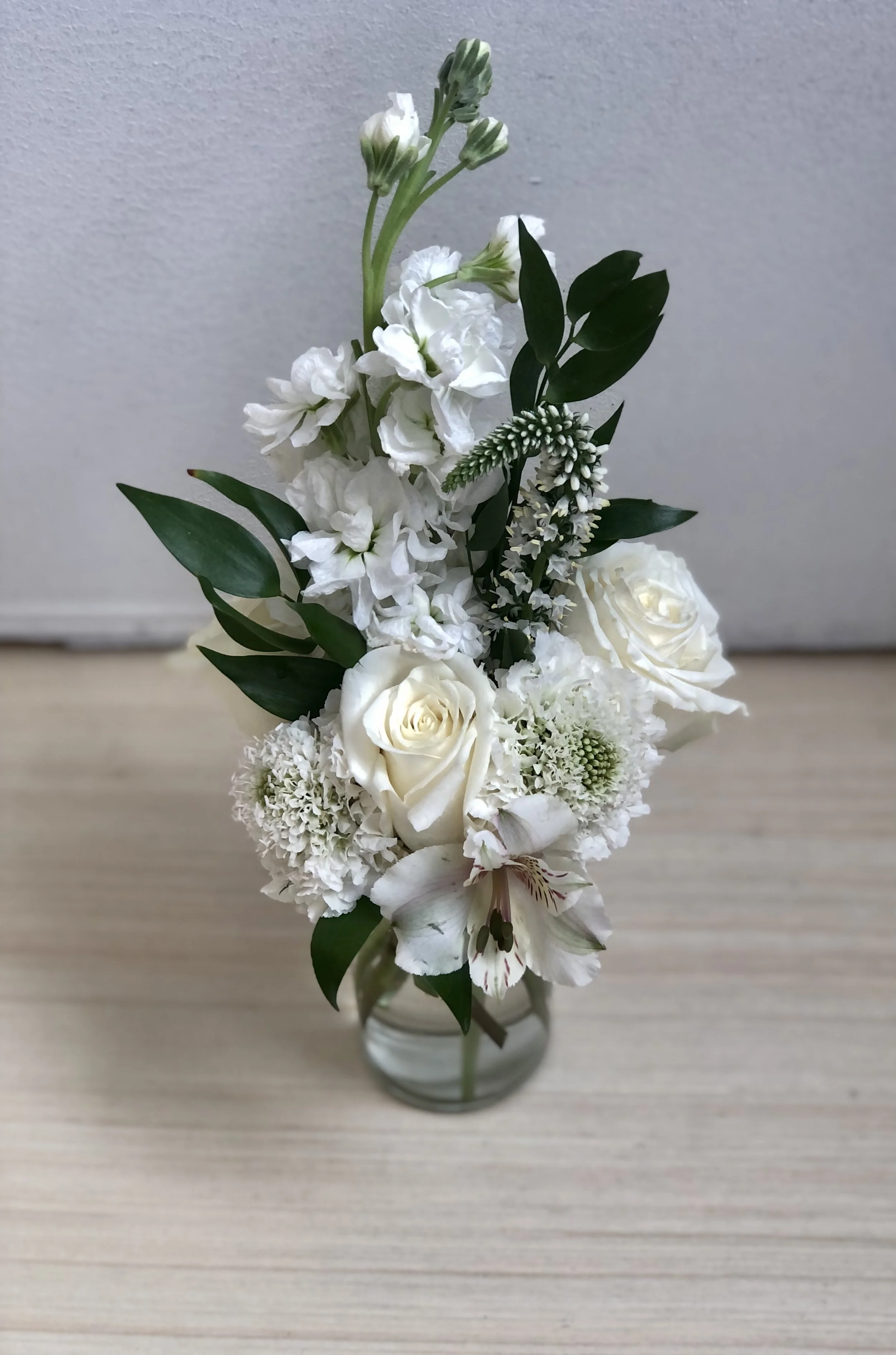 White floral arrangement in a clear glass vase on a light wooden surface against a gray background.