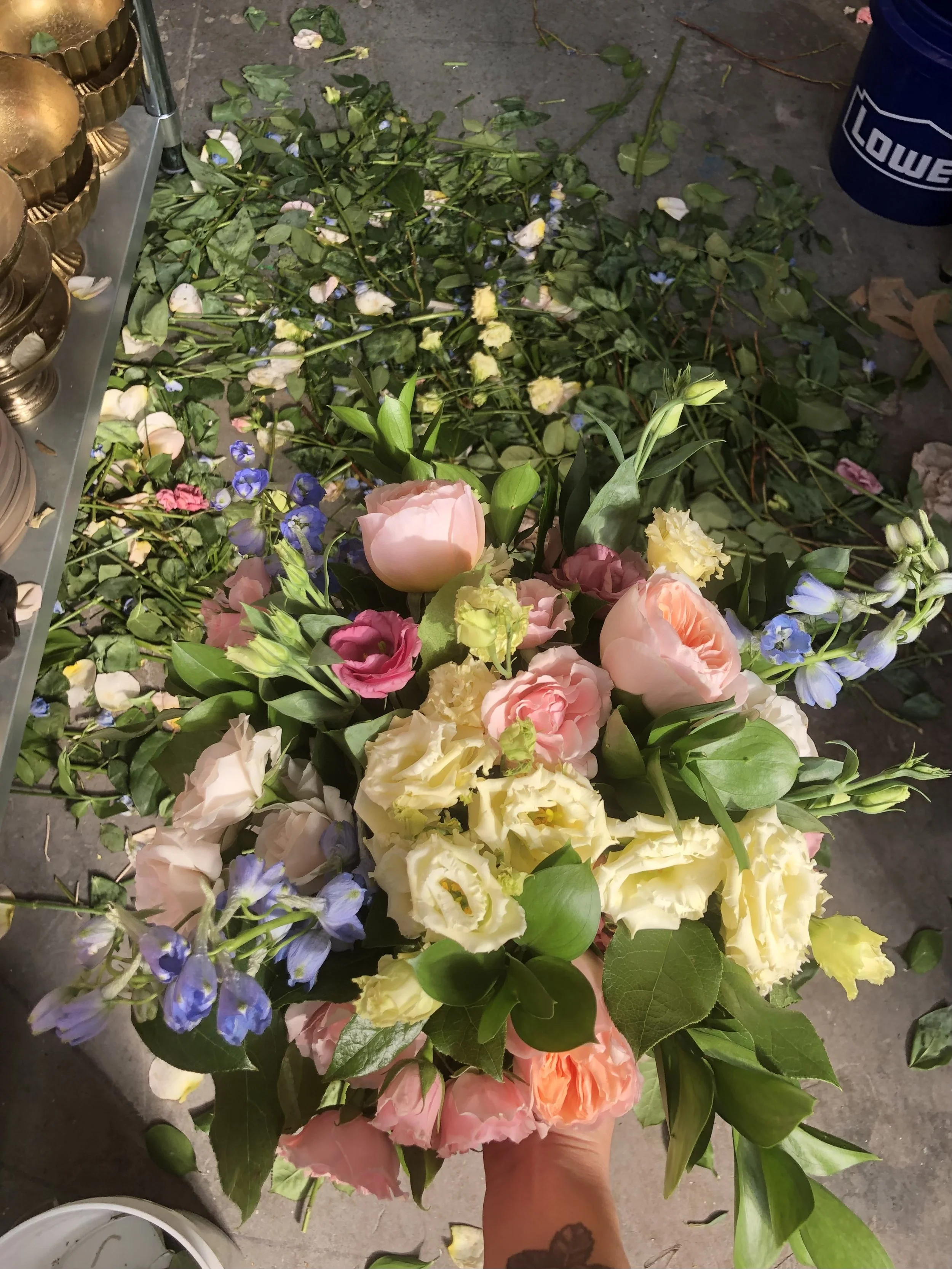 A person is holding a large colorful bouquet of flowers including pink tulips, yellow lisianthus, purple delphiniums, and other assorted blooms. Flower stems and leaves are scattered on the ground around them.