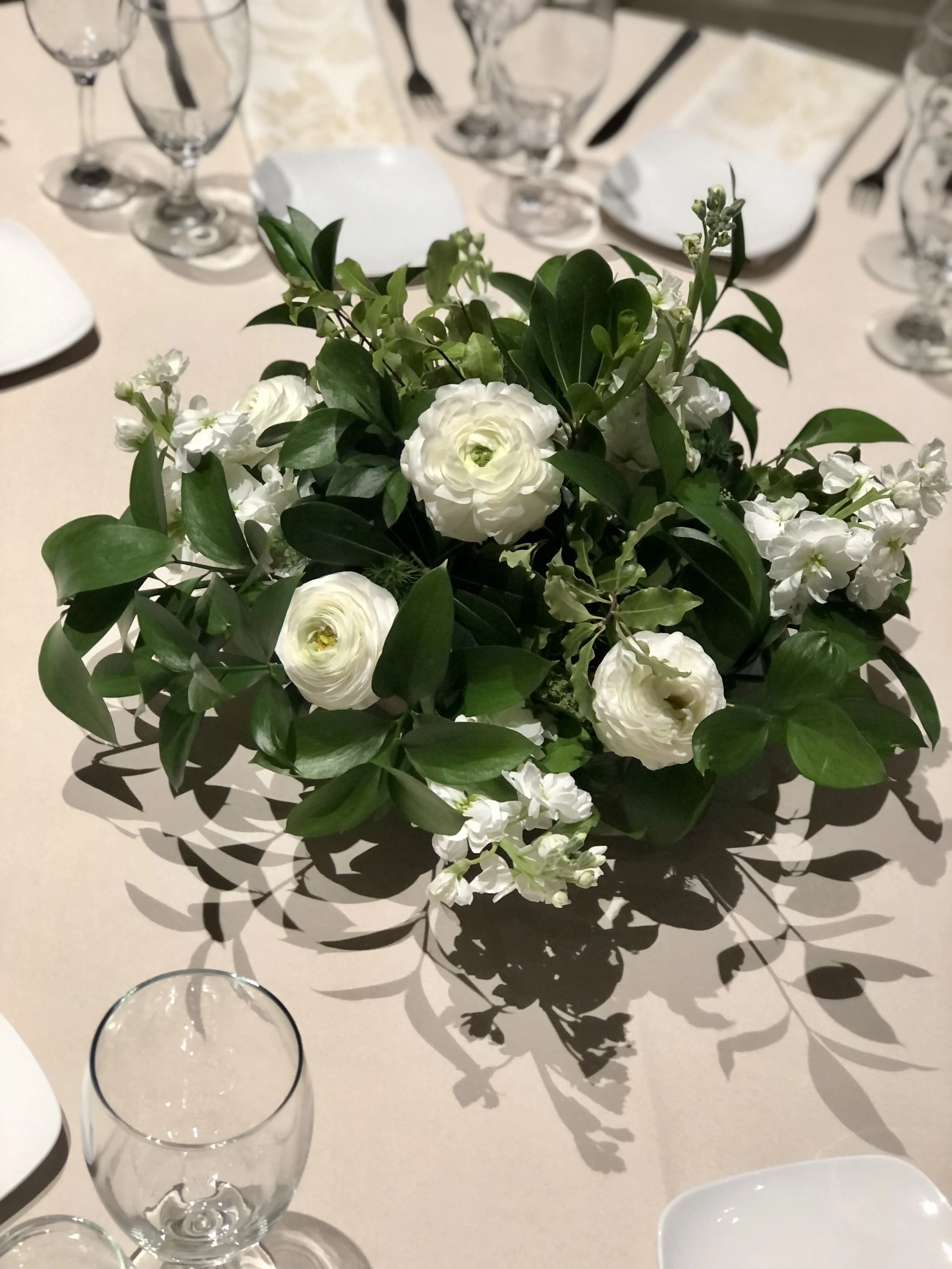 A centerpiece of white flowers and green foliage on a table with glassware and white plates.