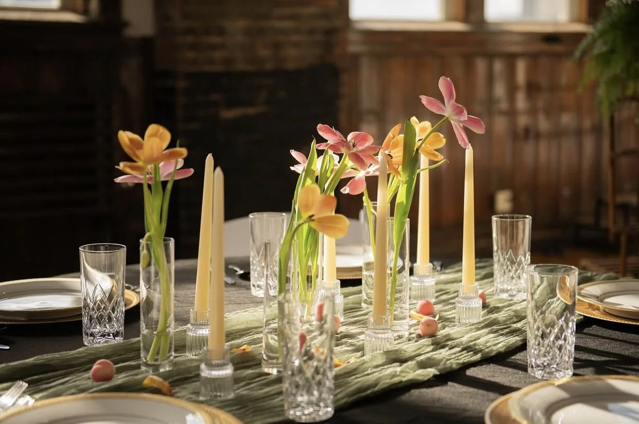 A beautifully set dining table decorated with pink and yellow flowers in glass vases, long taper candles, wine glasses, and plates with gold trim.