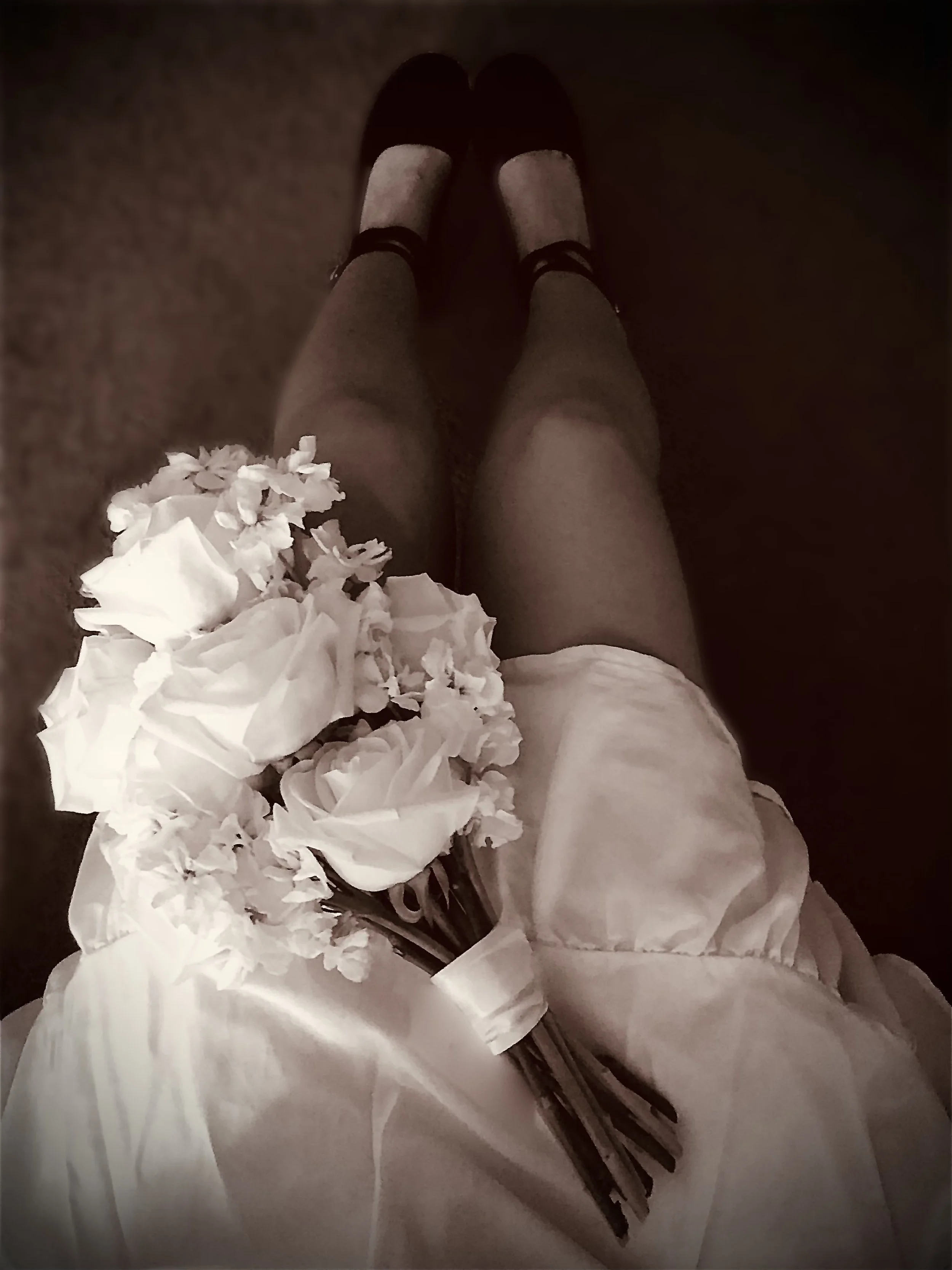 A person wearing black shoes, black socks, and a white dress, holding a bouquet of white flowers, sitting on a carpeted floor.