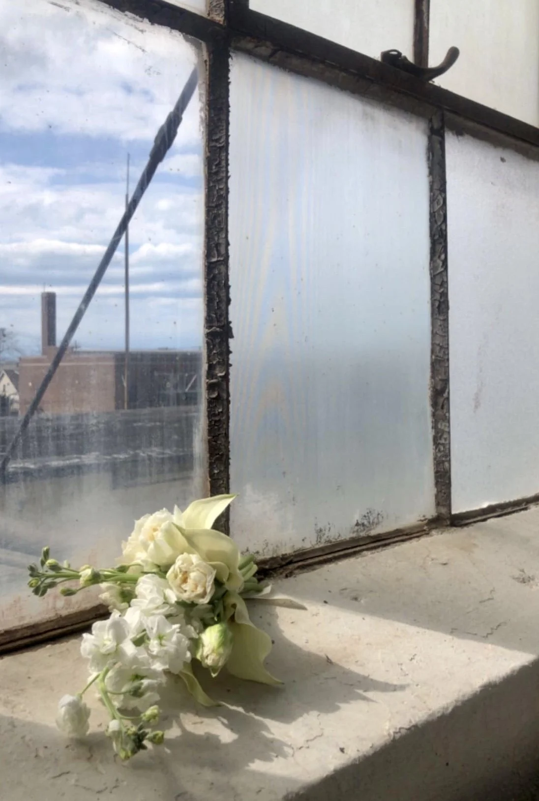 A small bouquet of white flowers resting on a windowsill next to a dirty, broken window with a cloudy sky outside.