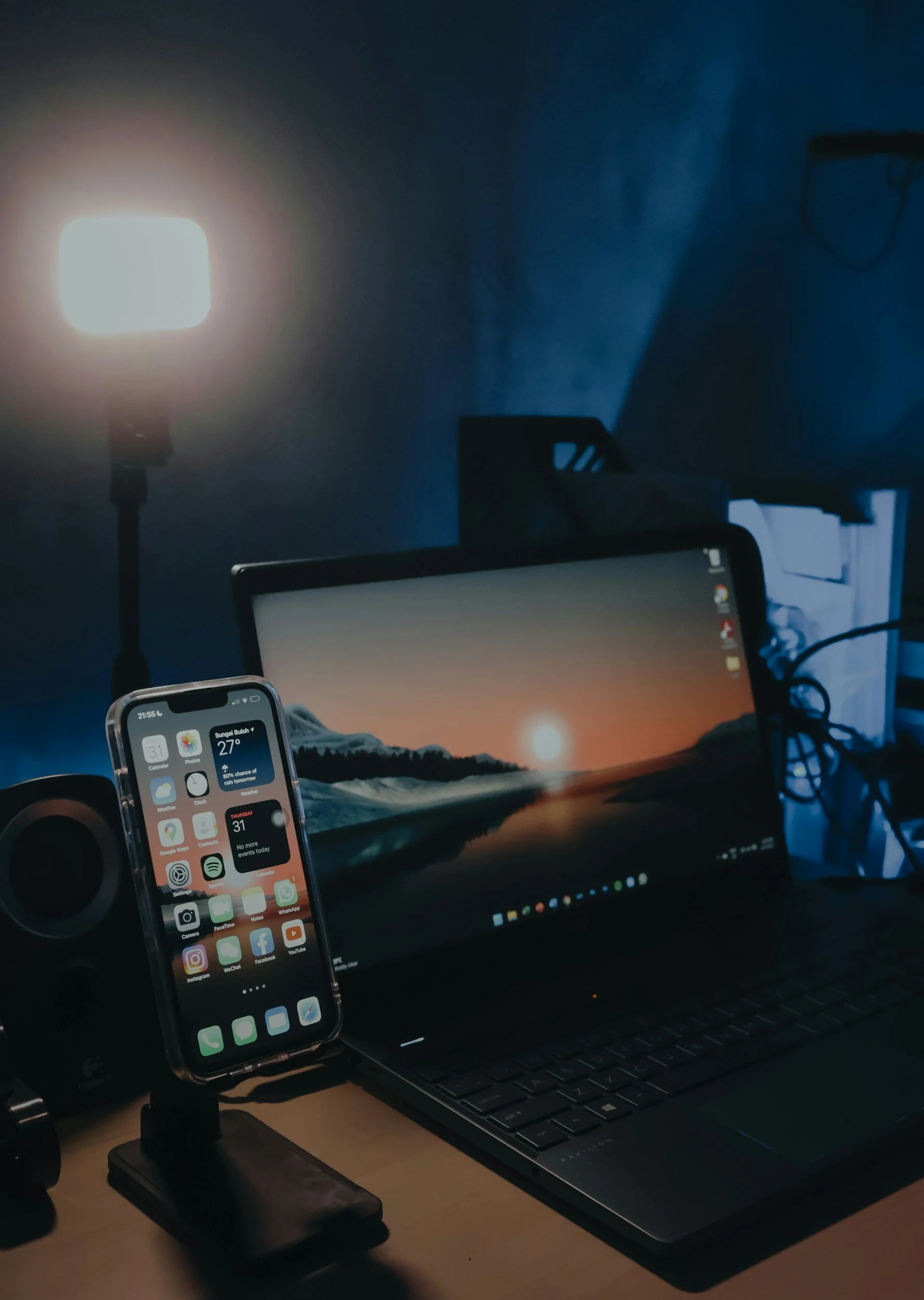 A workstation with a laptop showing a wave and sunrise wallpaper, a smartphone on a stand displaying app icons, a speaker, and a bright light in a dimly lit room.