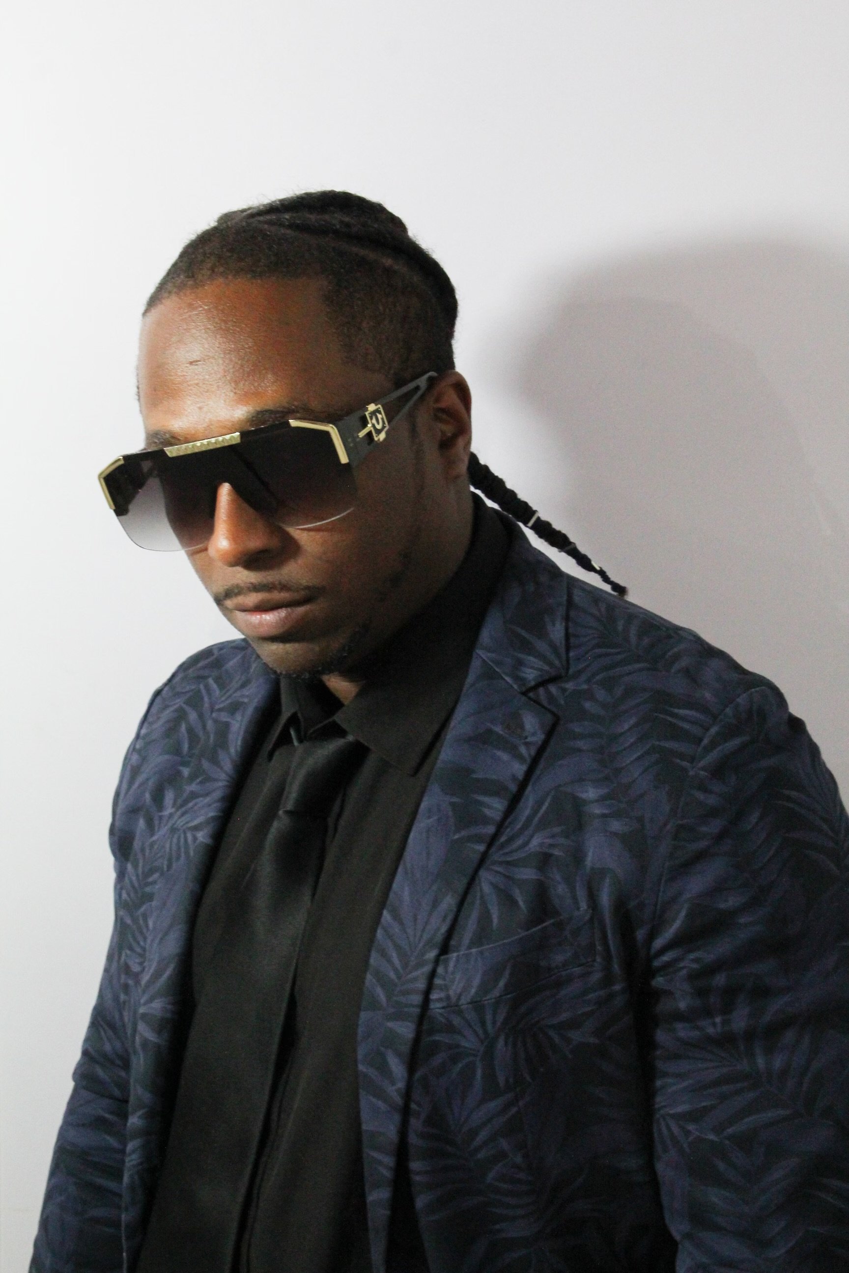 A man with dark skin wearing dark sunglasses, a black shirt, a dark patterned blazer, and a black tie, standing against a plain light-colored wall.