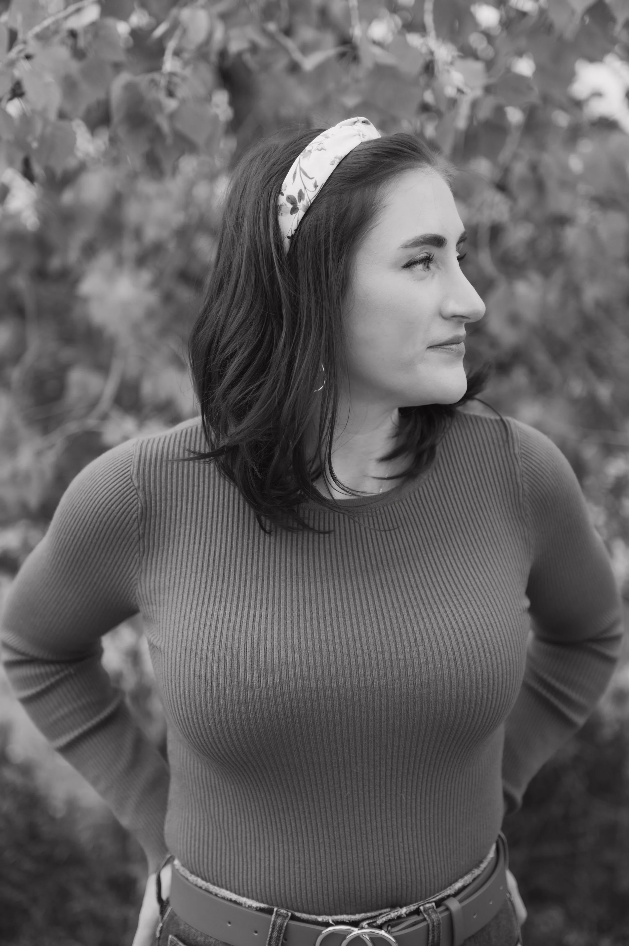 A woman with dark brown hair and blue eyes smiling outdoors, wearing an olive green long-sleeve top, blue jeans, a brown belt with gold rings, and a white headband, standing in front of a tree with yellow leaves.