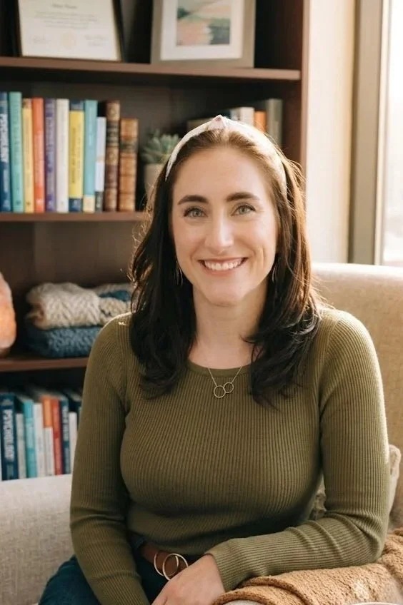 A woman with shoulder-length dark hair, wearing a headband and a green long-sleeve shirt, smiling while sitting on a beige armchair in a room with a bookshelf behind her filled with books and decorative items. Sunlight is streaming in through a window.